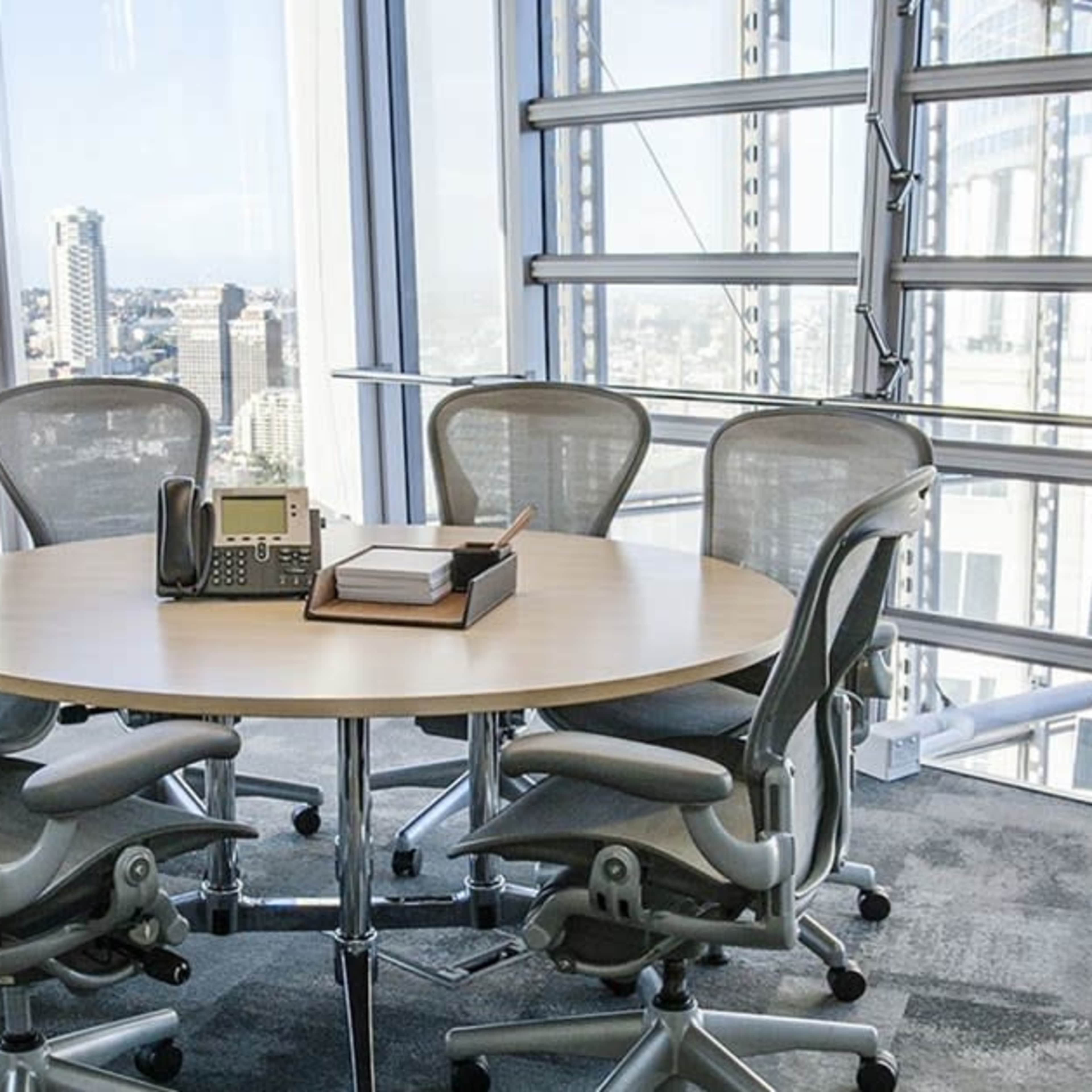 A modern conference room features a round table surrounded by ergonomic chairs with large windows offering a city view.