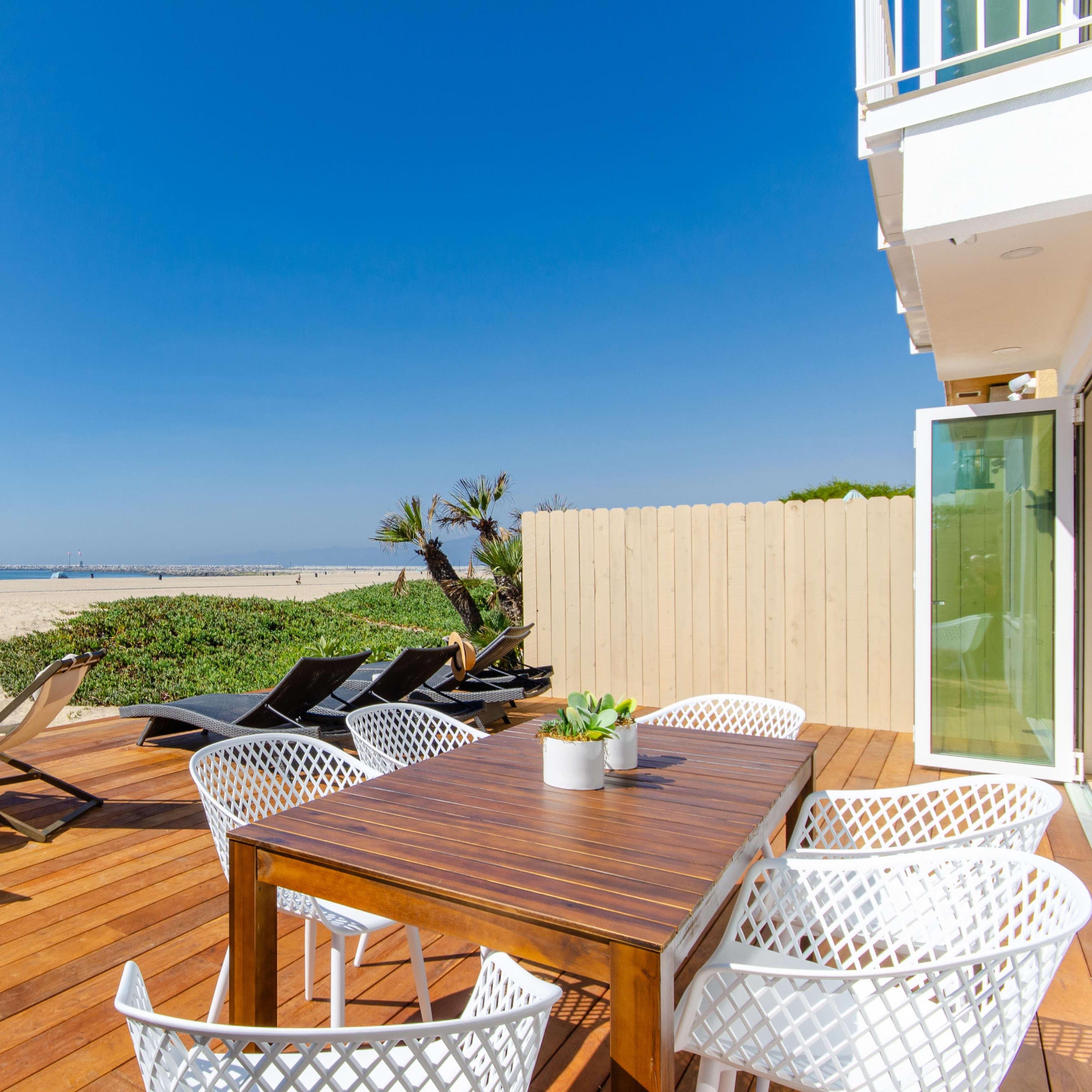 A wooden deck with a table and chairs overlooks a sandy beach against a clear blue sky.