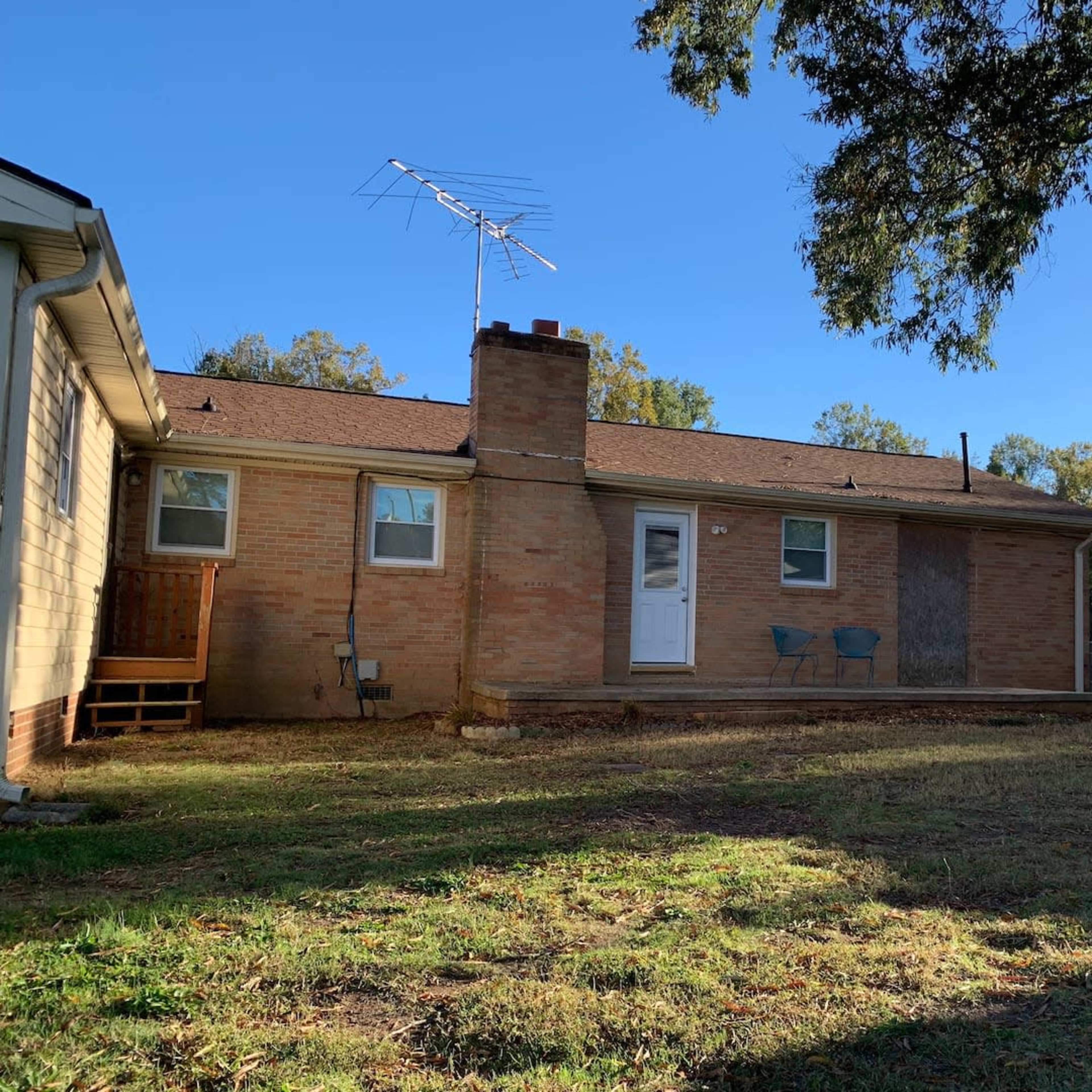 A brick house with a small porch, two blue chairs, and a chimney, surrounded by a grassy yard.
