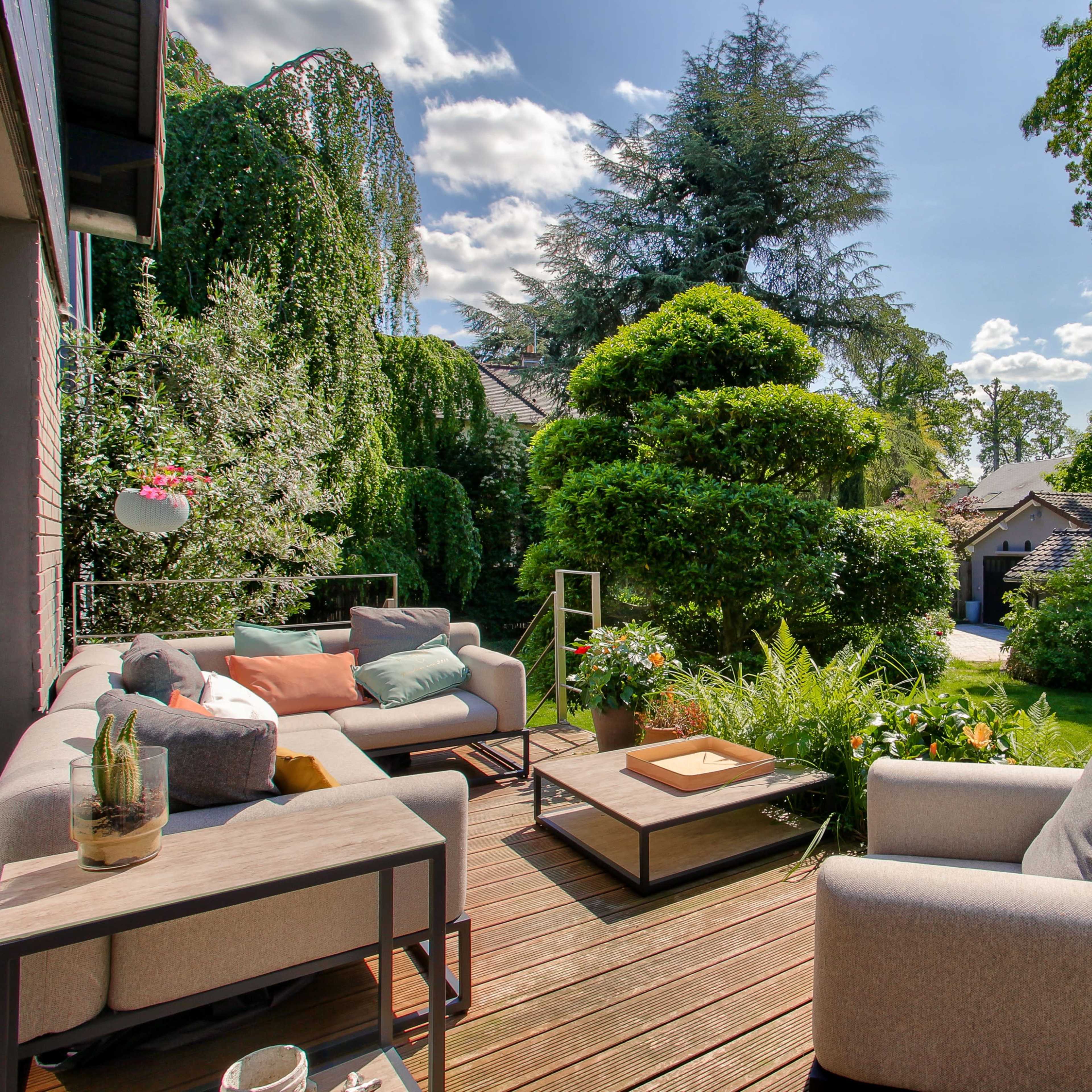 A spacious outdoor seating area with modern furniture overlooks a lush garden filled with trees and colorful plants.
