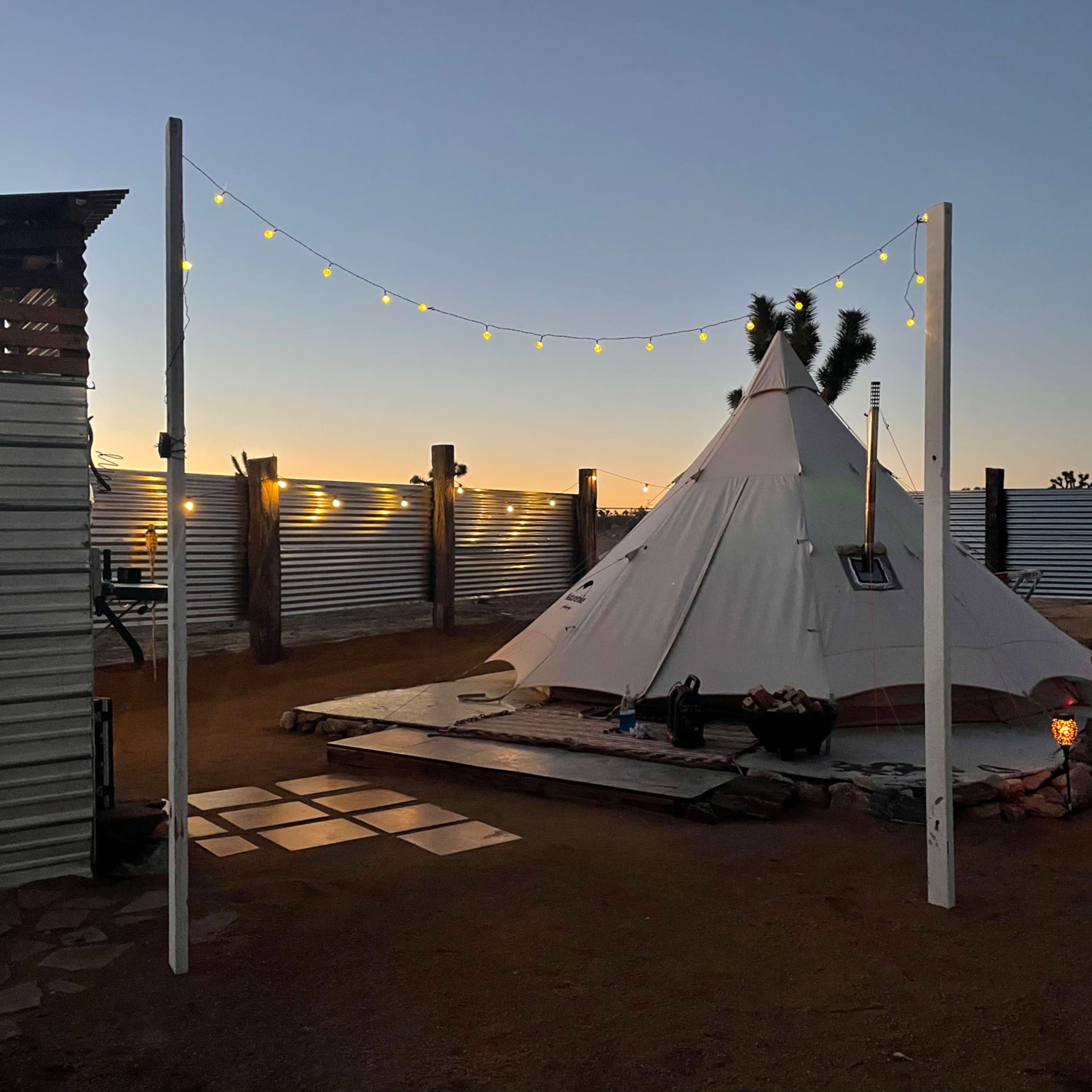 A teepee sits on a wooden platform surrounded by decorative lights and stone pathways in a desert setting at dusk.