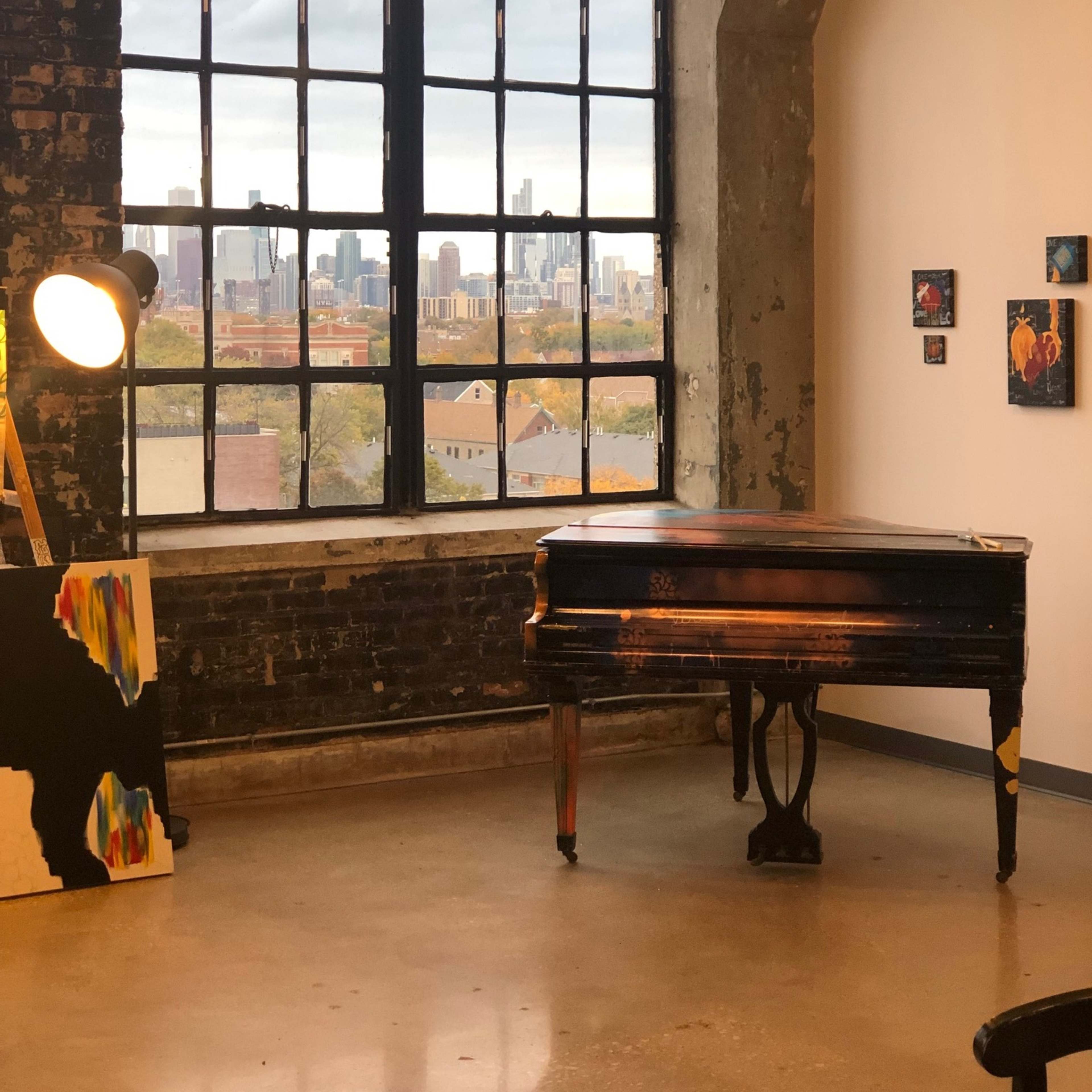 A grand piano is positioned in front of a large window overlooking a city skyline, with artwork and a standing light nearby.