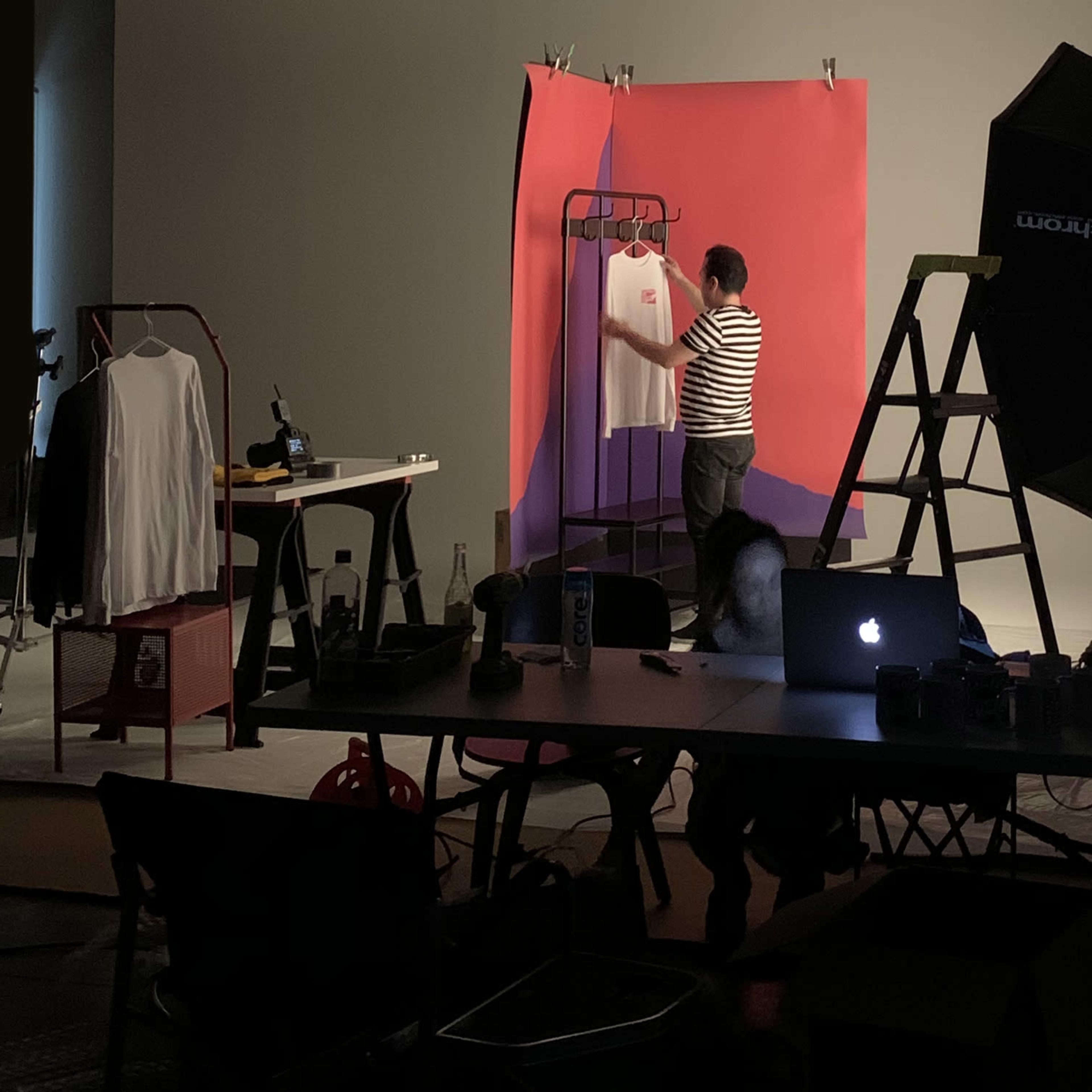 A person in a striped shirt adjusts a garment on a rack in a photography studio with colorful backdrops and equipment.