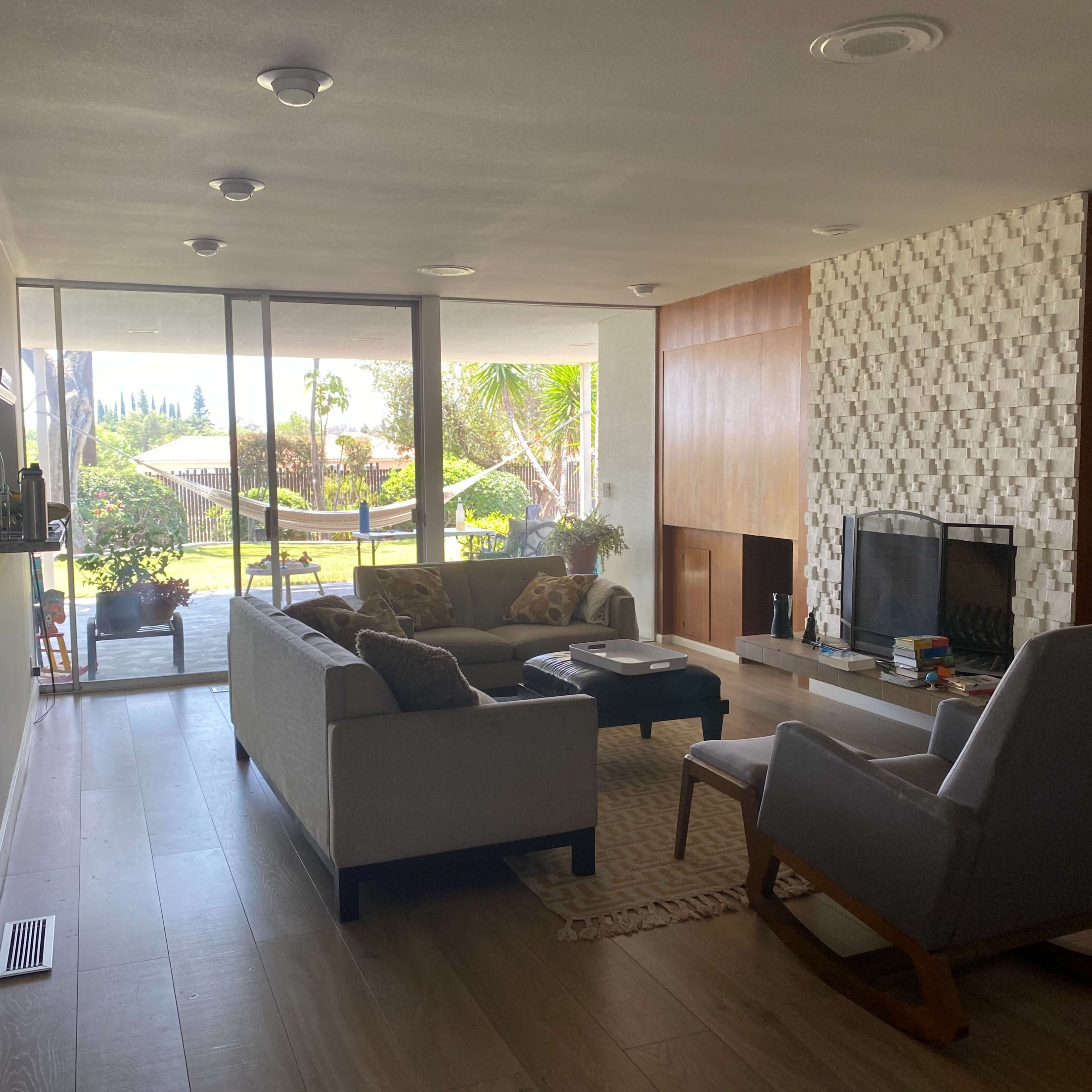 A living room features a sofa, an armchair, and a fireplace, with large windows overlooking a backyard garden.