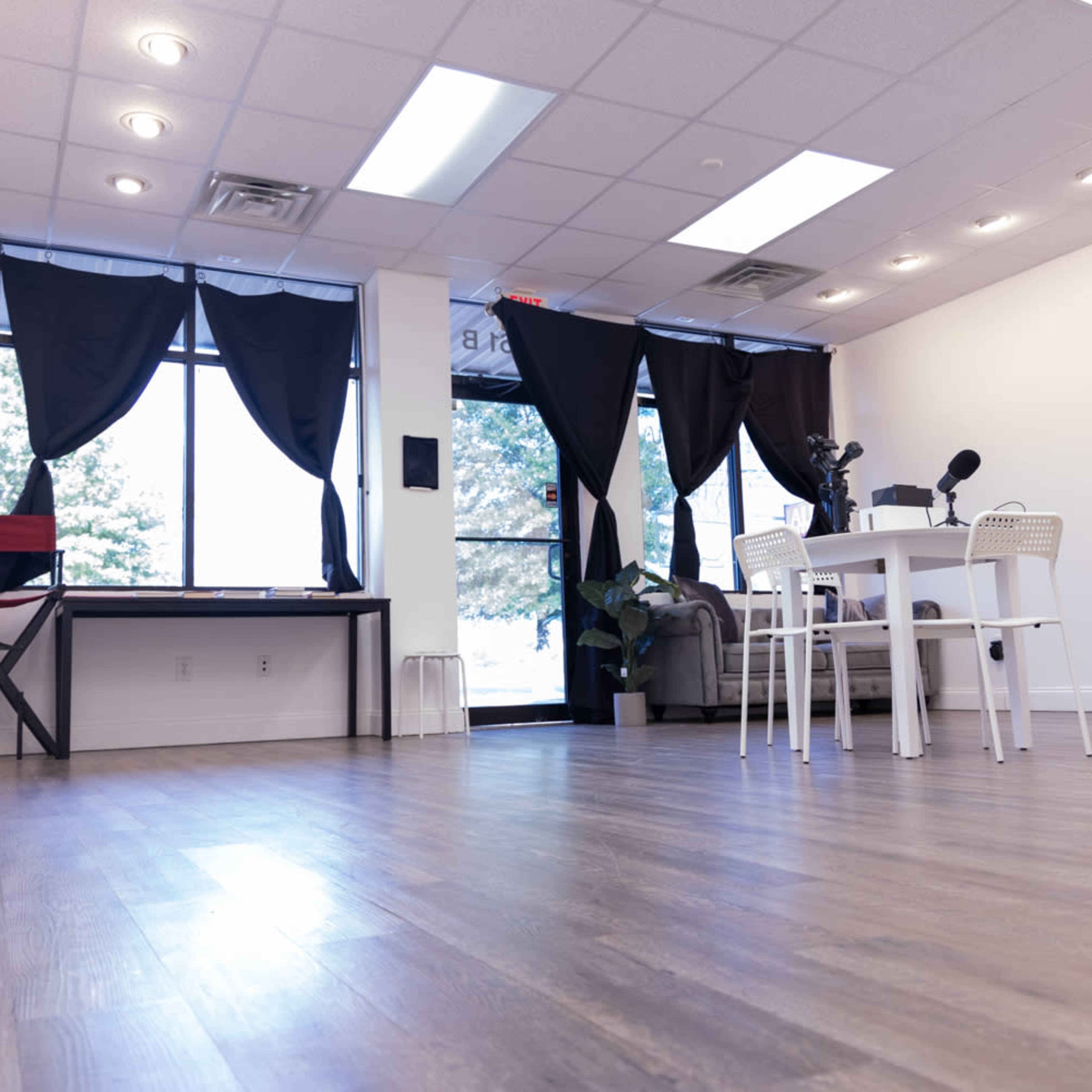 A bright, spacious room with a table and chairs, a desk, and large windows covered by black curtains.