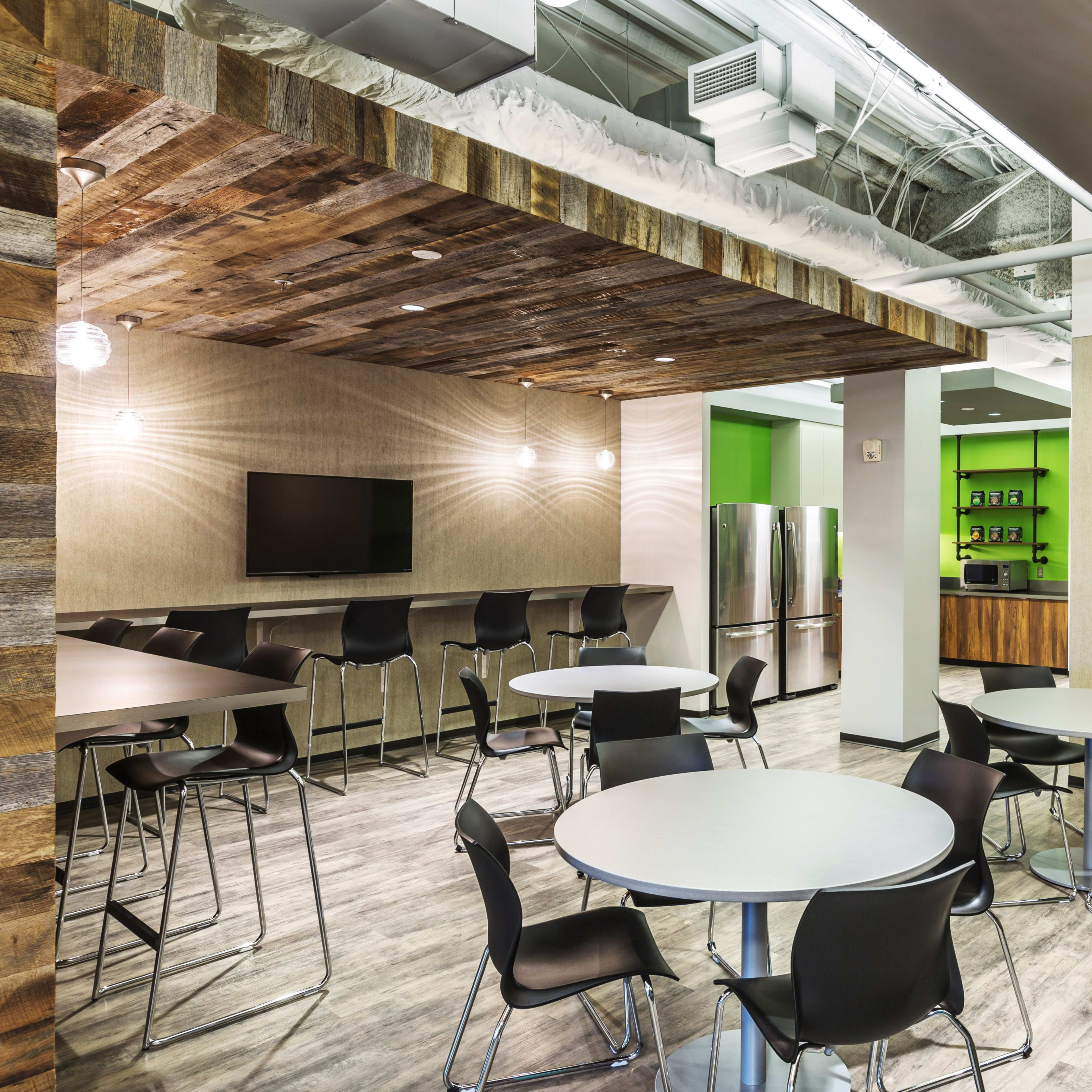 A modern communal kitchen area with tables, chairs, a television, and a kitchen counter featuring an open layout and wooden accents.