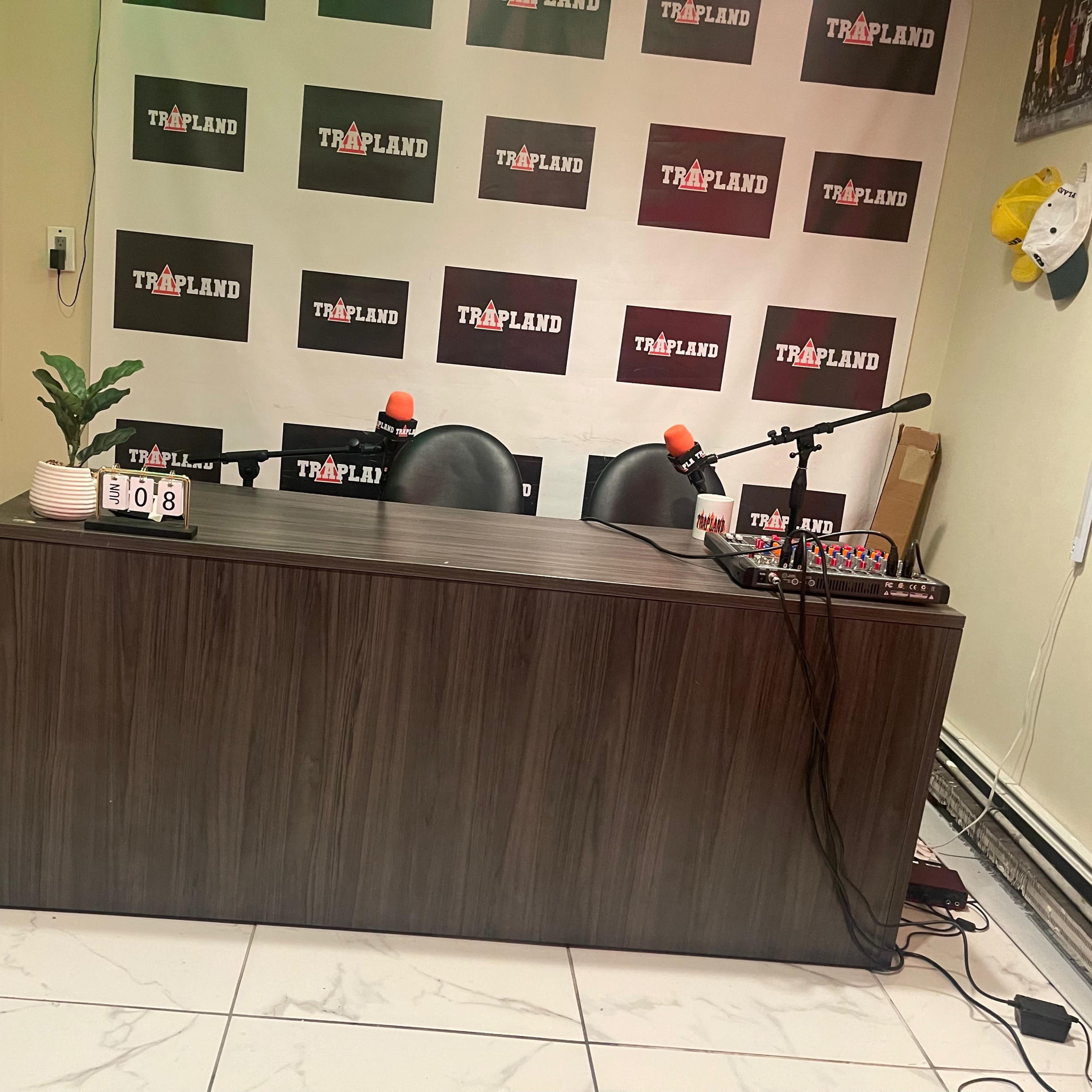 A recording studio setup with a wooden table, two chairs, and microphones, against a backdrop of branded posters.
