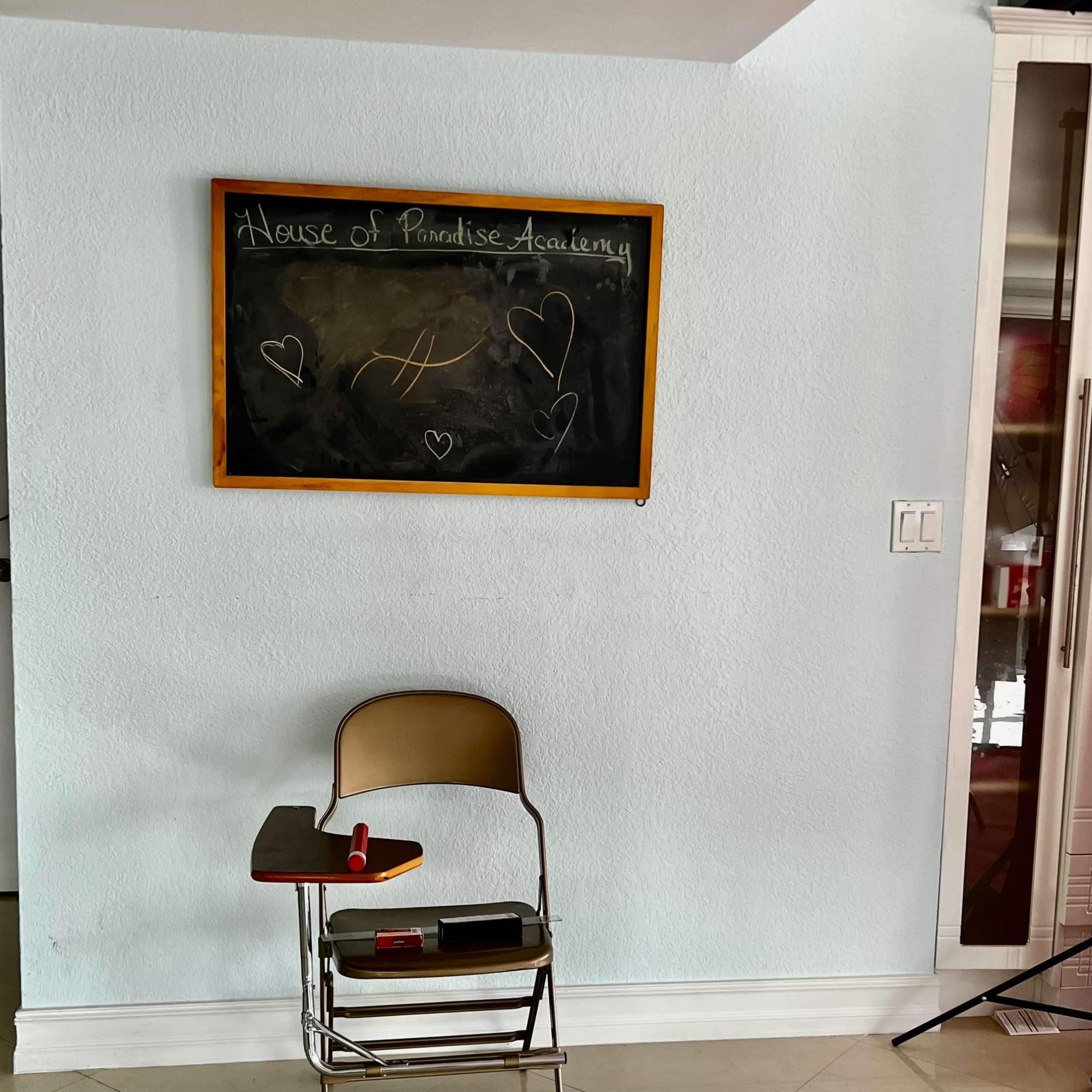 A classroom scene with a single metal desk and a chalkboard that reads "House of Paradise Academy" on a light blue wall.