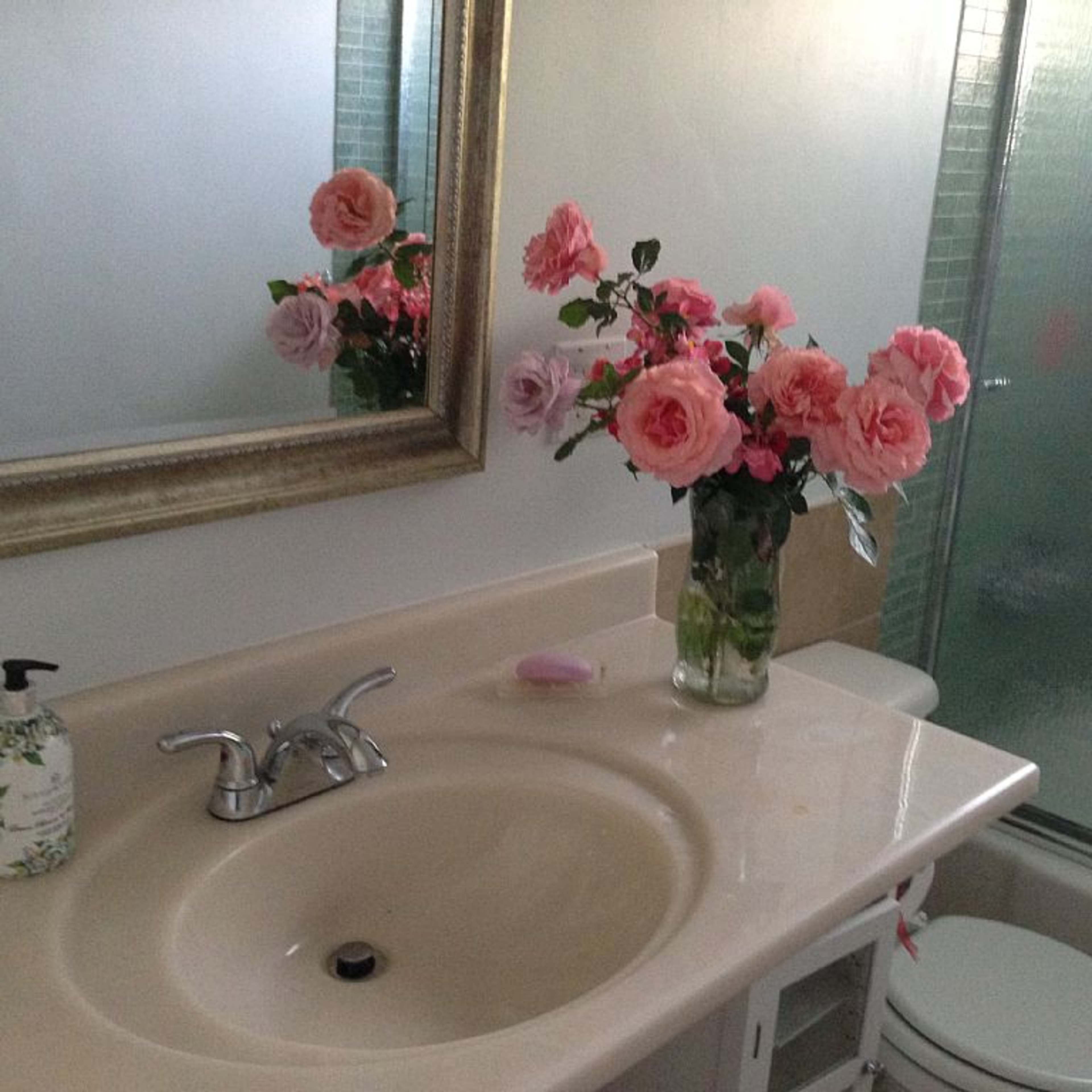 A bathroom features a sink with a decorative soap dispenser, a large mirror, and a vase of pink and purple roses on the countertop.
