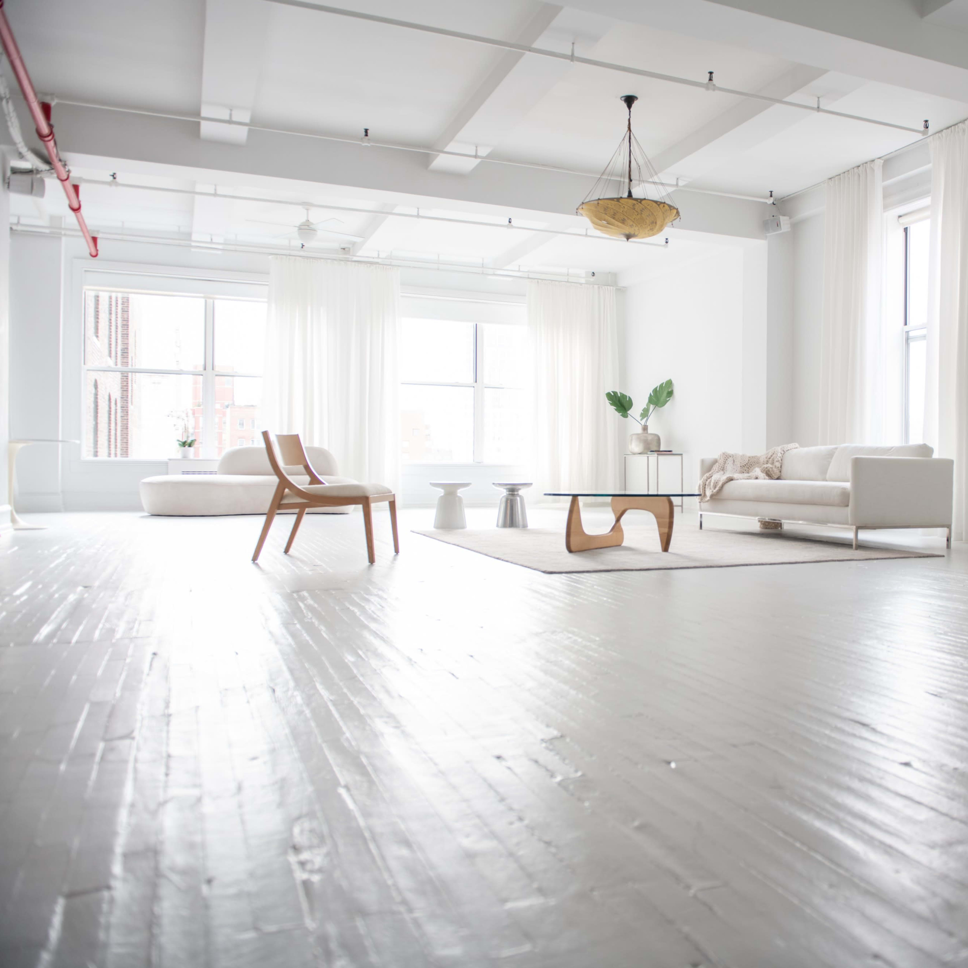 A spacious, white-walled room features minimalist furniture, large windows with sheer curtains, and a light wooden floor.