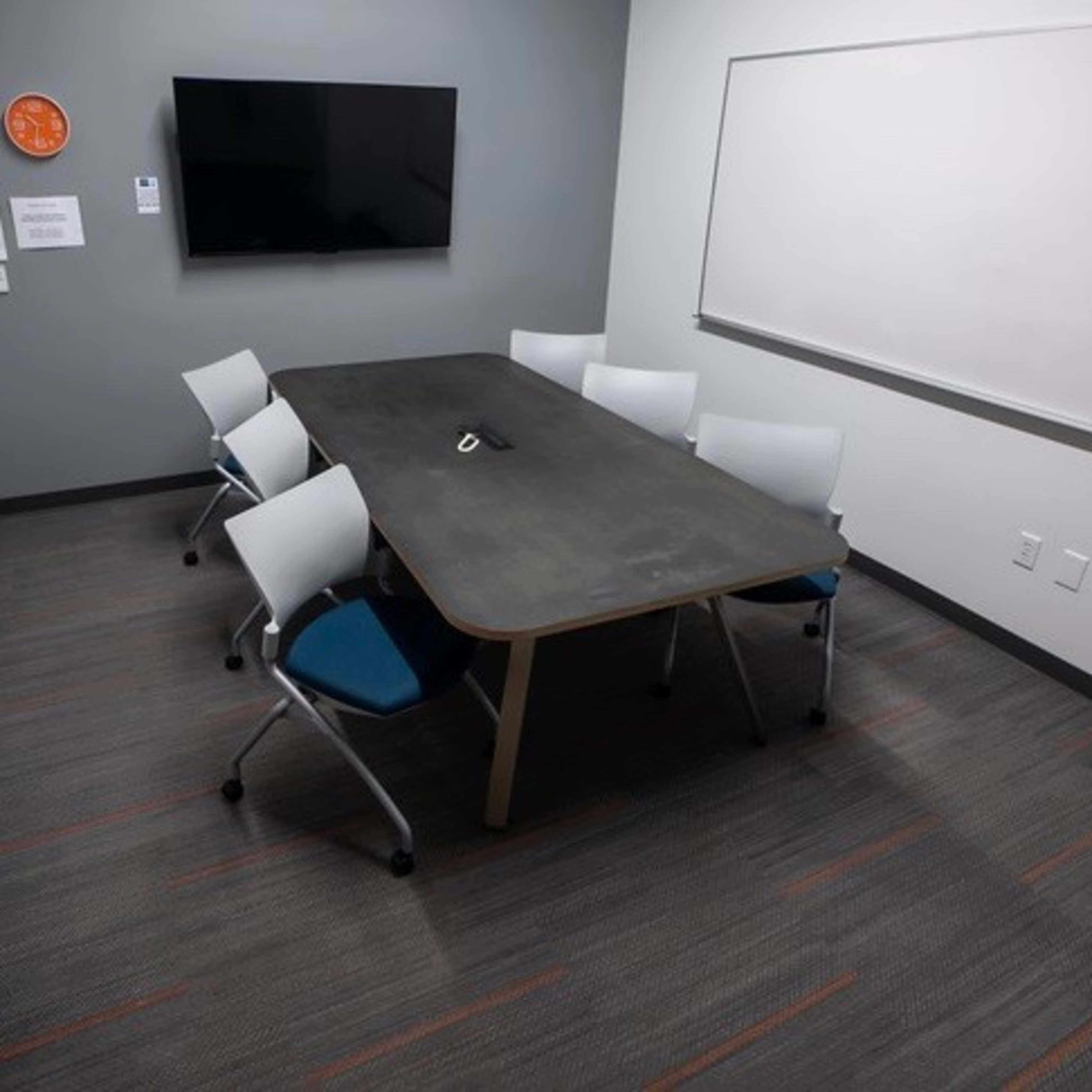 A conference room with a large table surrounded by six chairs, a wall-mounted television, and a whiteboard.