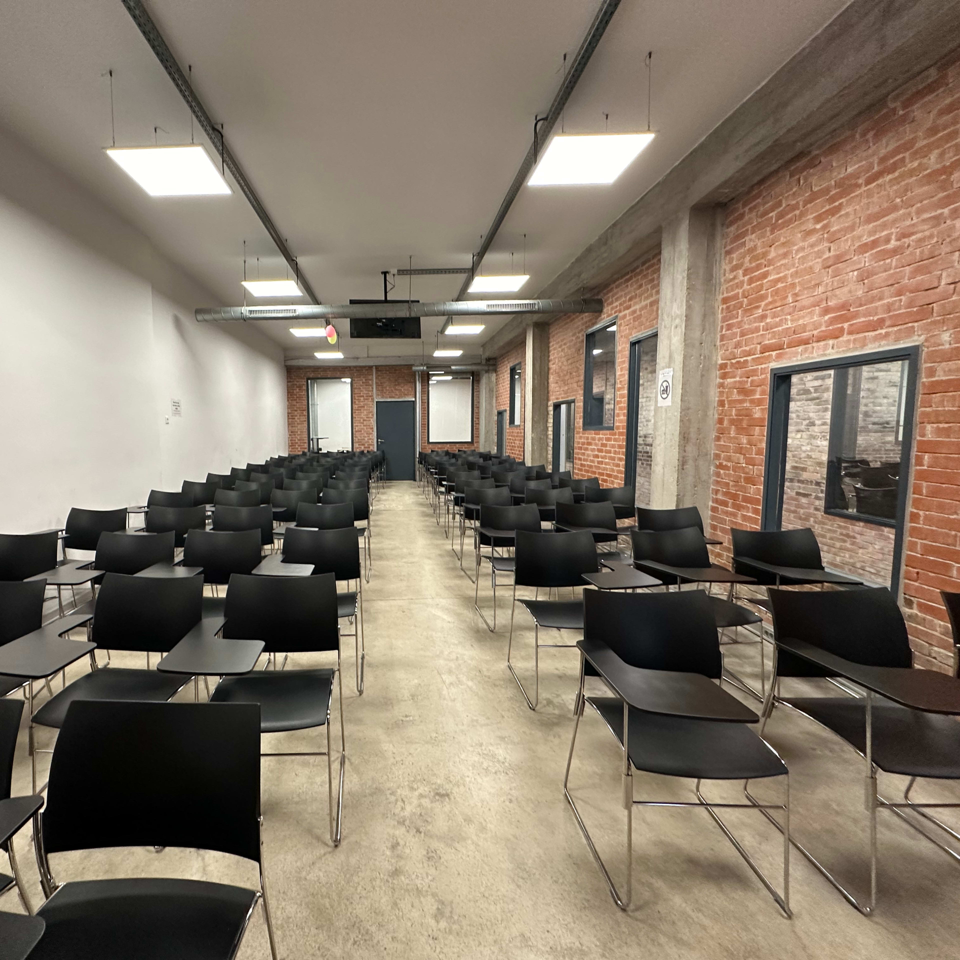 An empty classroom arranged with numerous black chairs in rows facing a front wall.