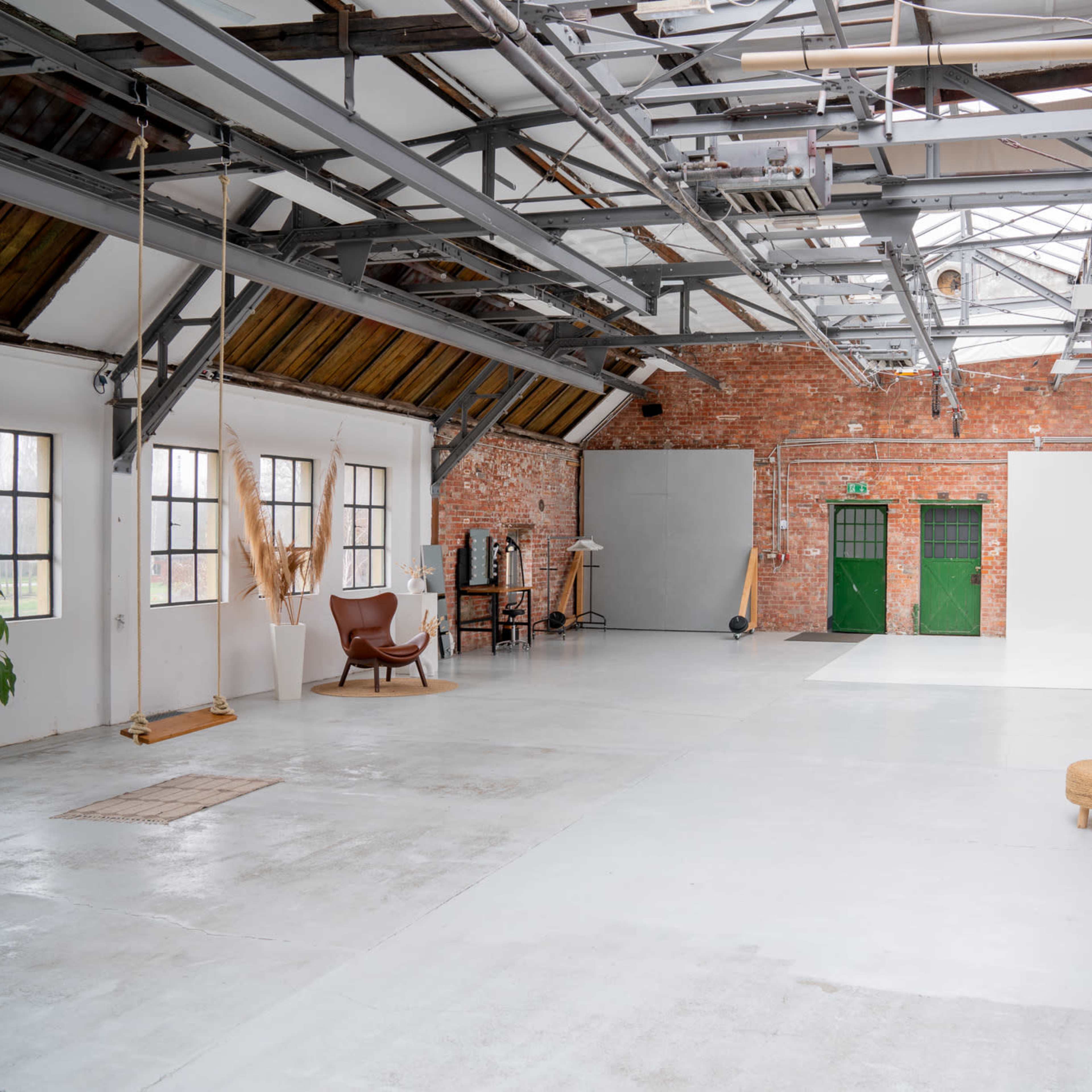 A spacious, industrial-style studio with exposed brick walls, high ceilings, and various furniture arrangements, including a sofa and a stool.