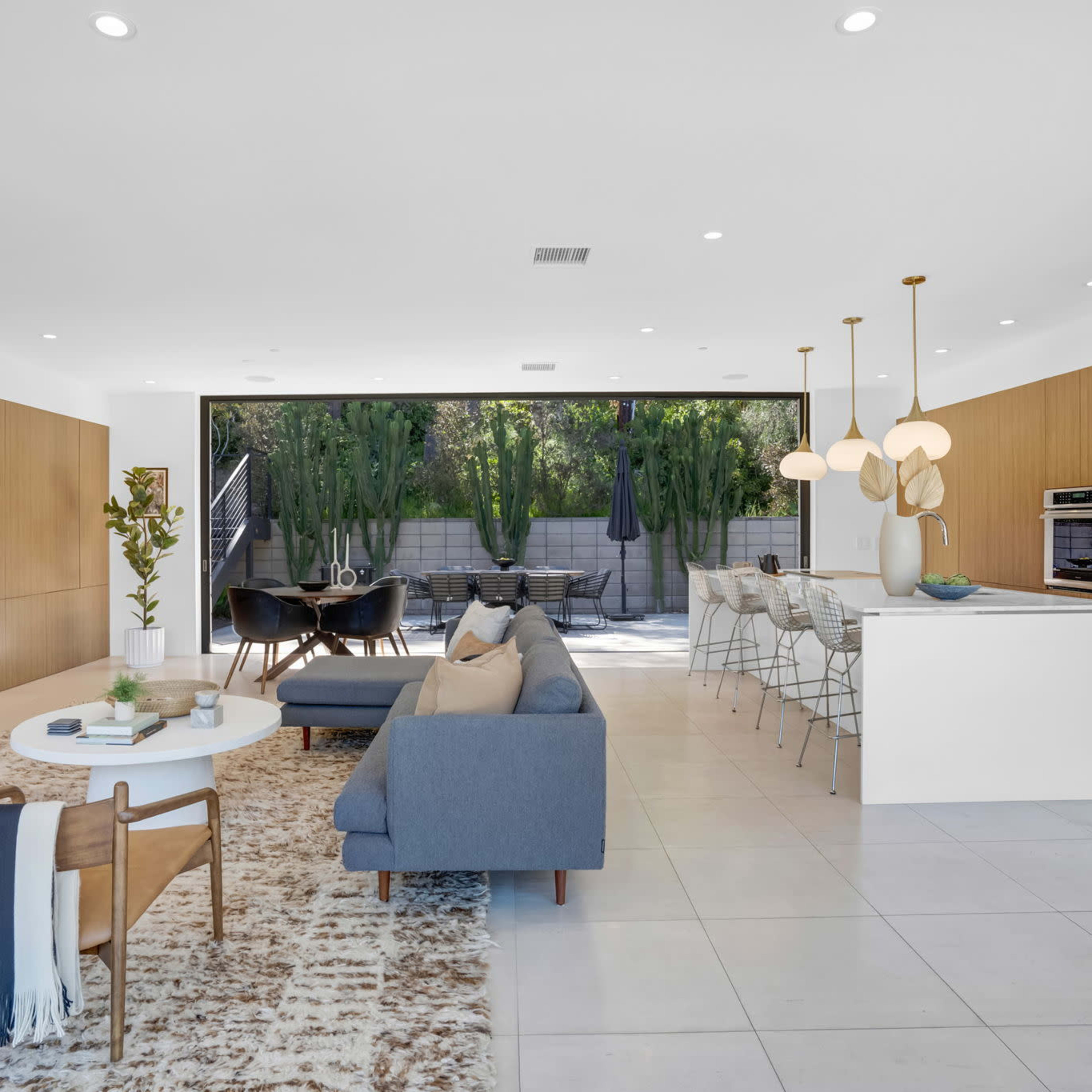 A modern living space with a fireplace, a seating area, and an open kitchen that leads to an outdoor patio surrounded by greenery.