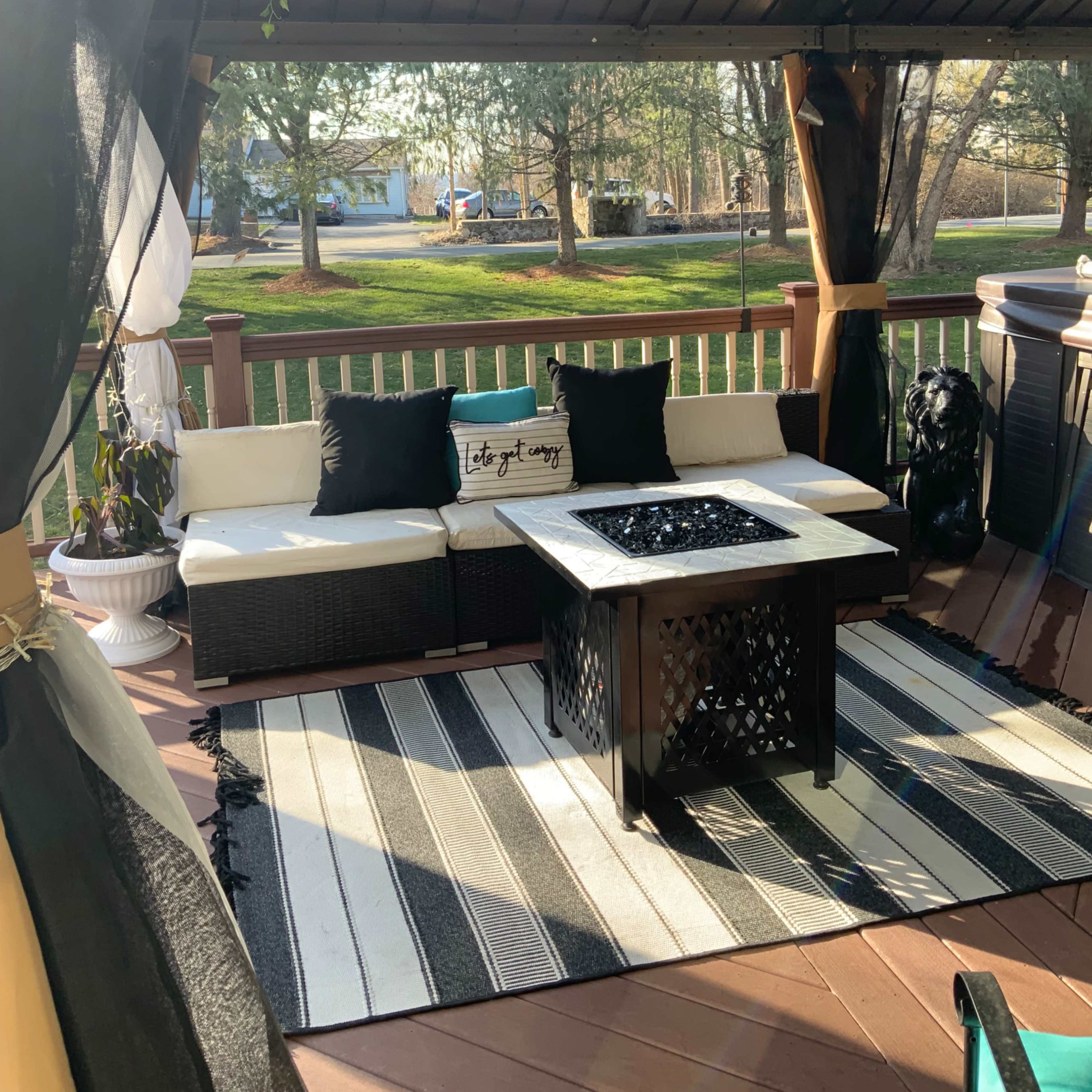 The scene shows a gazebo with a seating area featuring a sectional couch, a coffee table with a fire pit, and a striped rug, surrounded by trees in a yard.