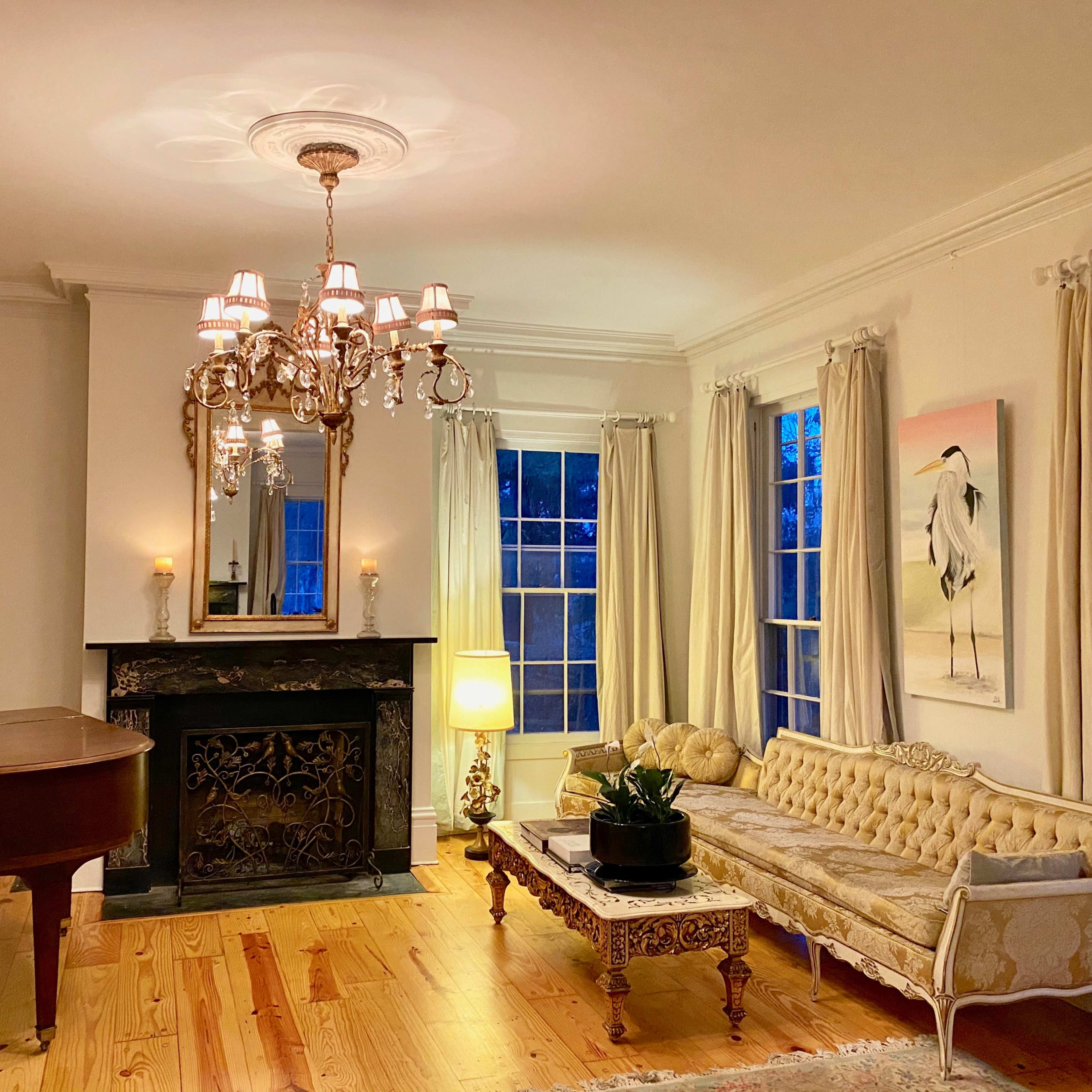 A elegantly furnished living room with a chandelier, a grand piano, a floral sofa, a decorative fireplace, and large windows allowing natural light.