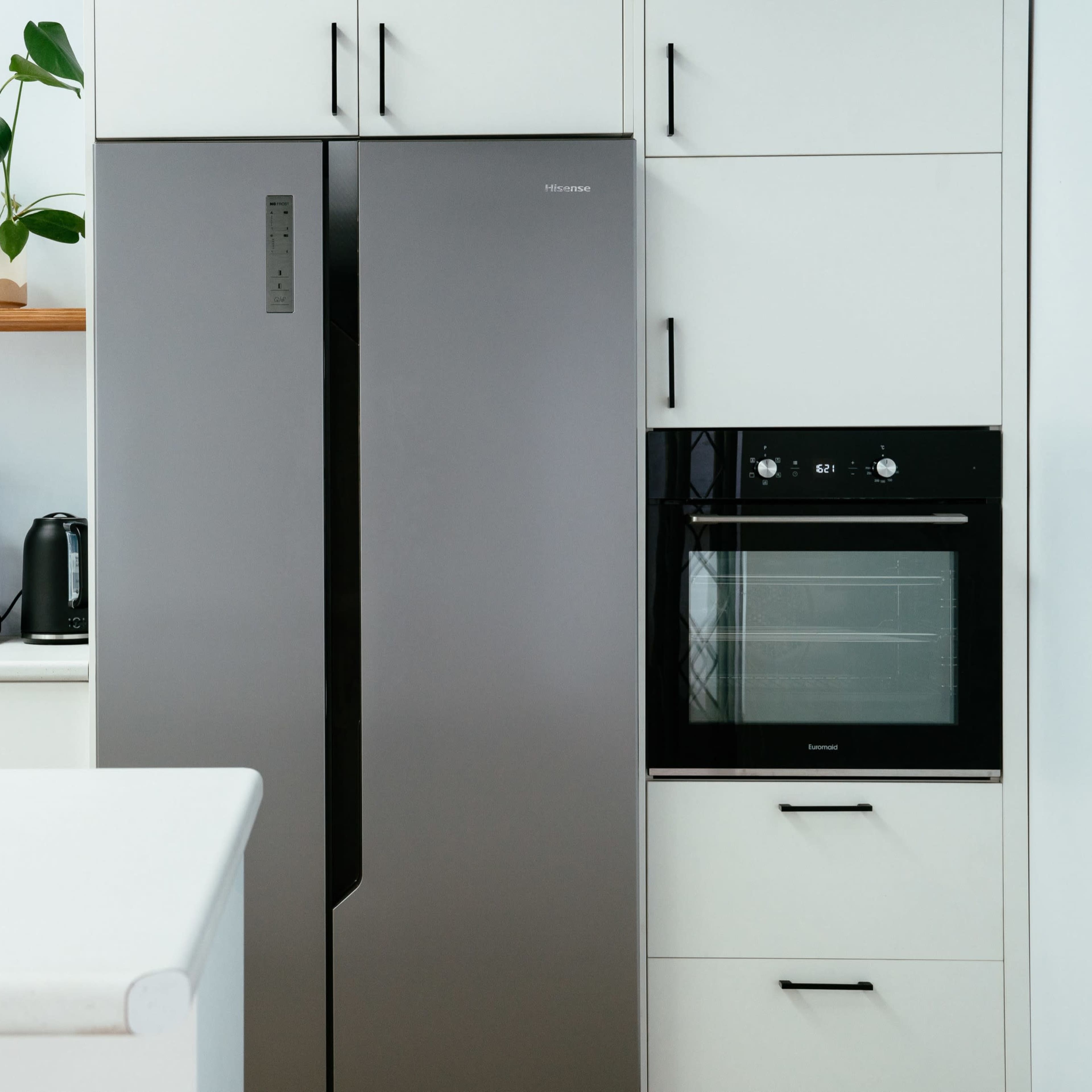 A modern kitchen featuring a silver refrigerator, an oven, and a coffee maker on a counter, with potted plants on a shelf.