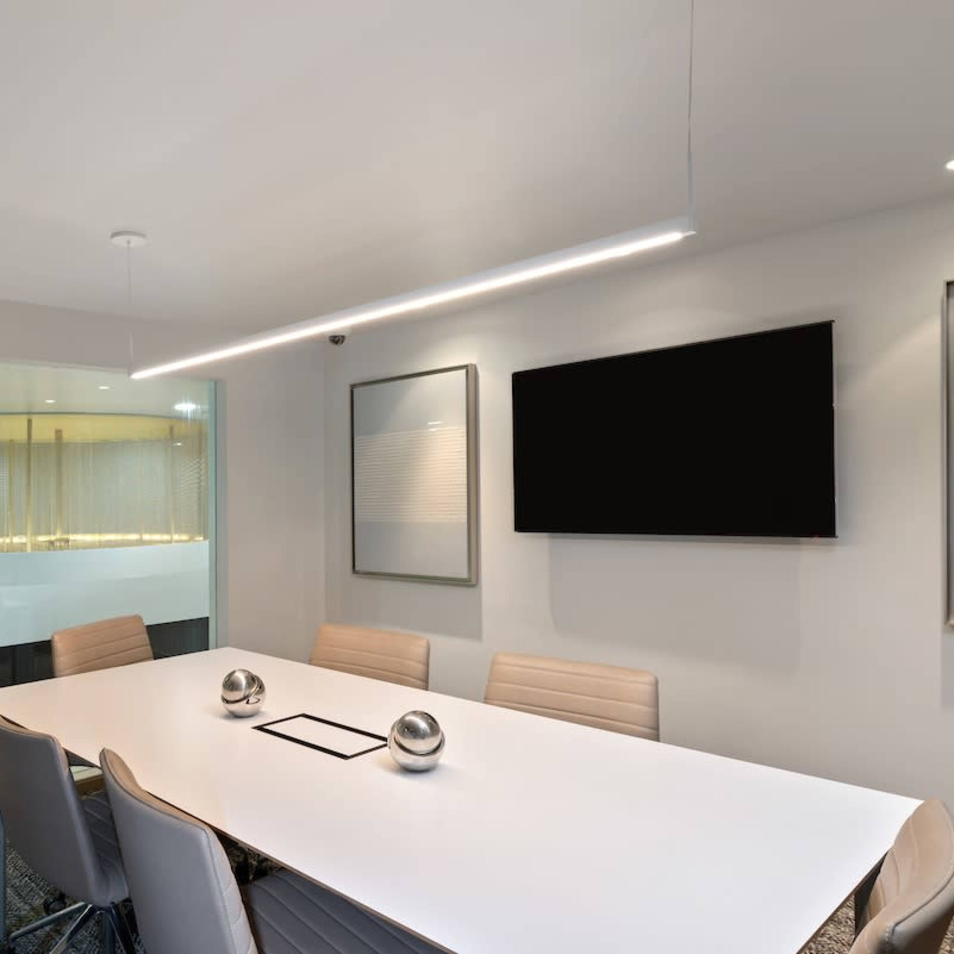 A modern conference room features a large white table surrounded by gray chairs, with a wall-mounted television and a glass partition leading to another space.