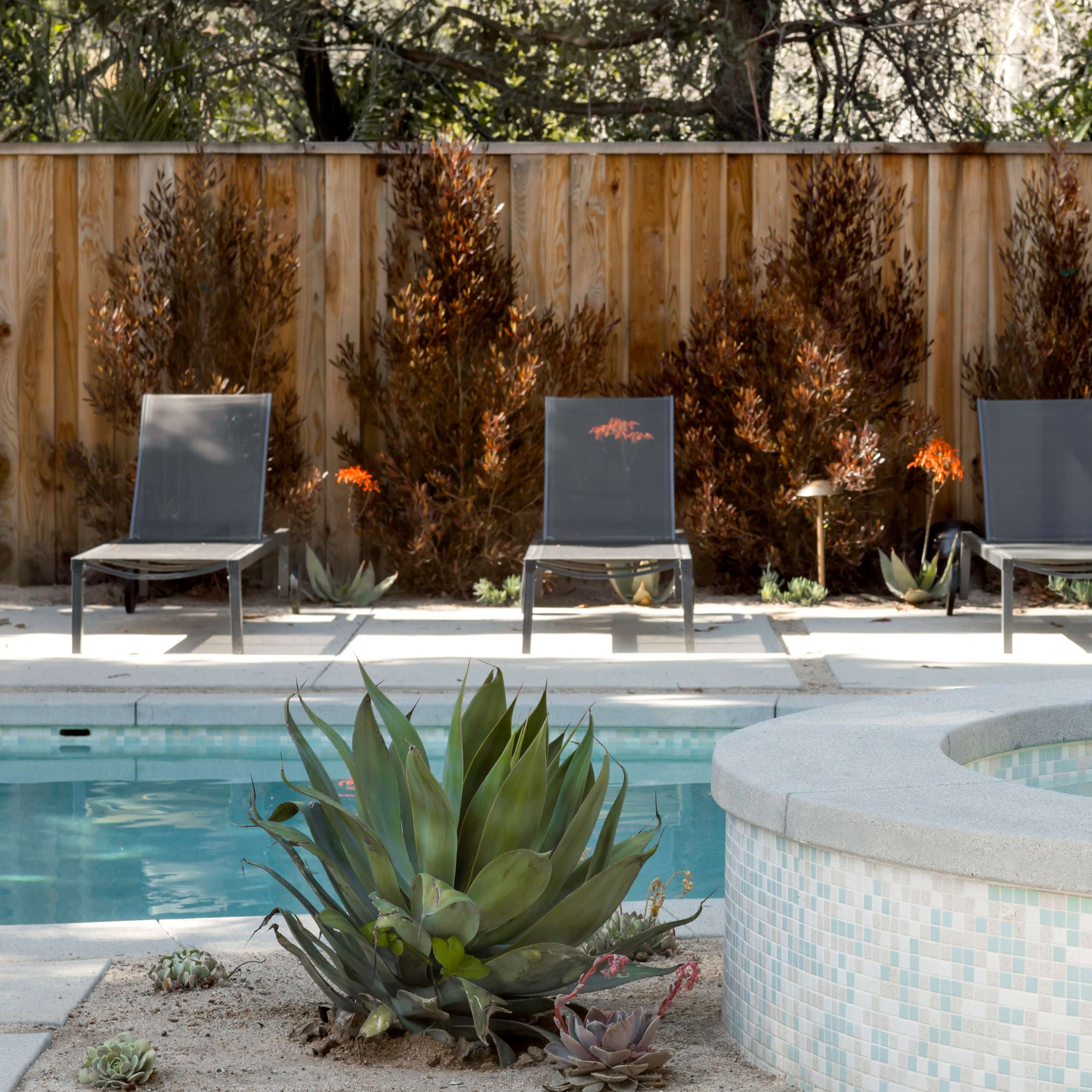 A backyard pool area featuring four lounge chairs, a circular hot tub, and landscaped greenery along a wooden fence.