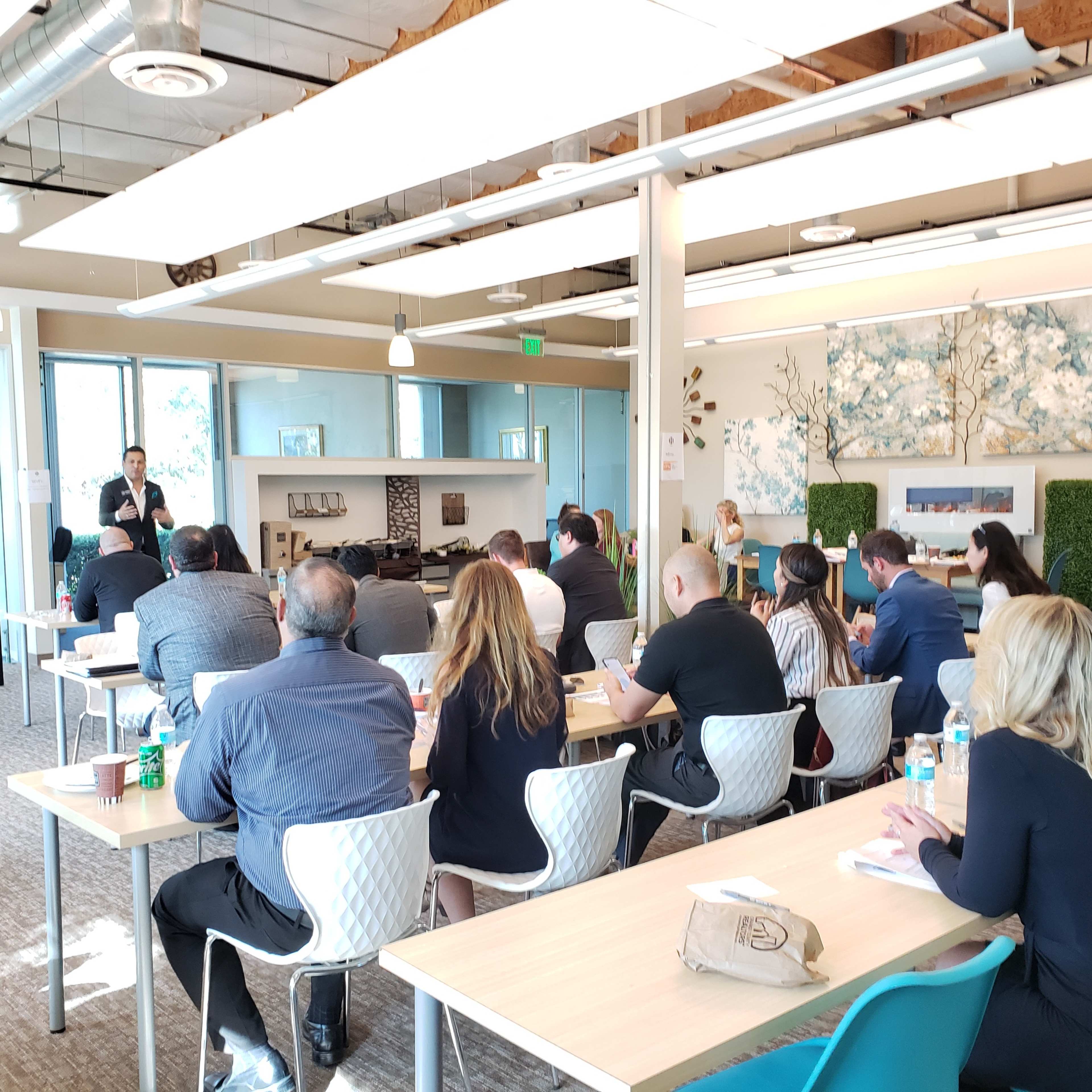 A speaker presents to an audience seated at tables in a modern conference room.