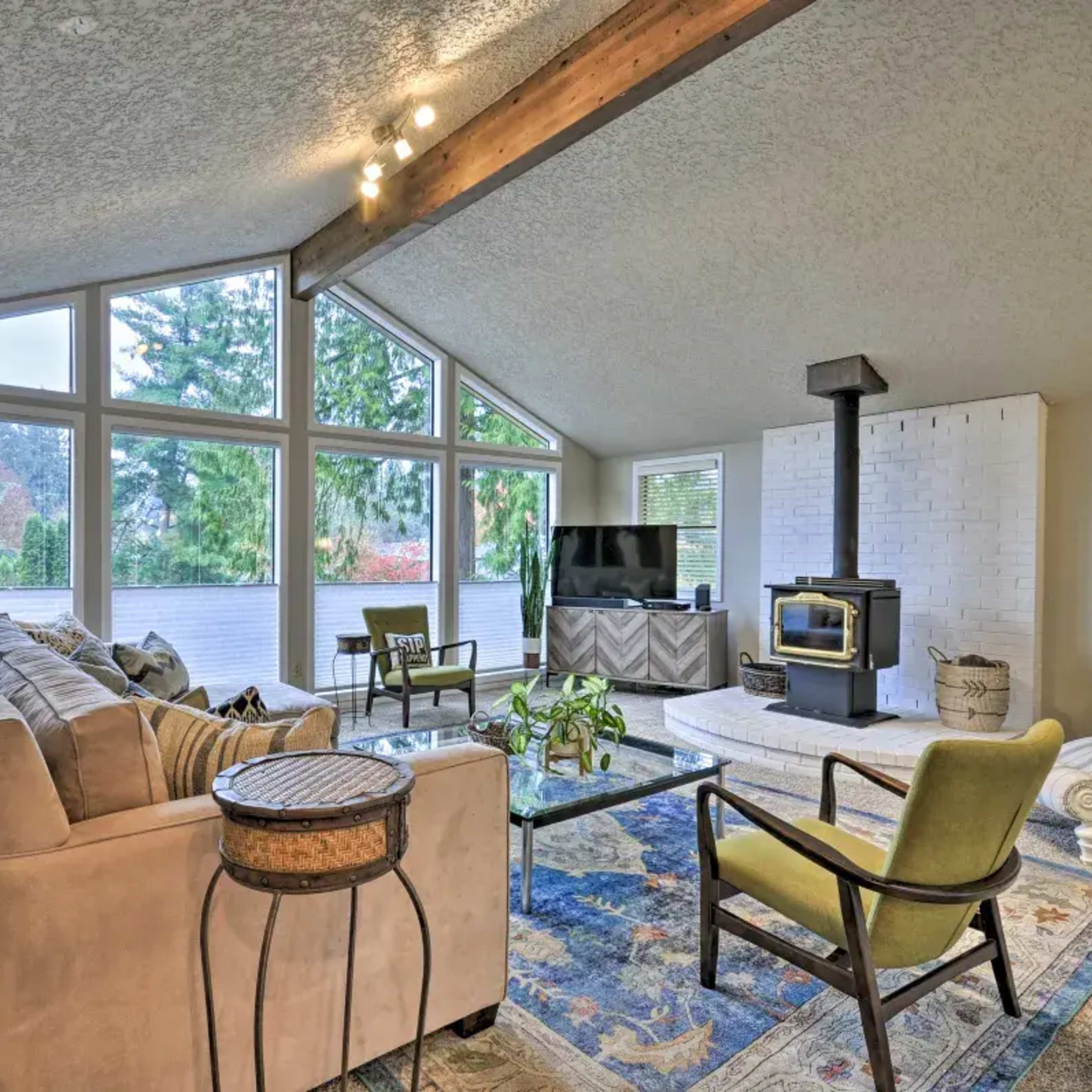 A cozy living room with a high ceiling, large windows, a wood stove, and modern furniture arranged around a patterned rug.