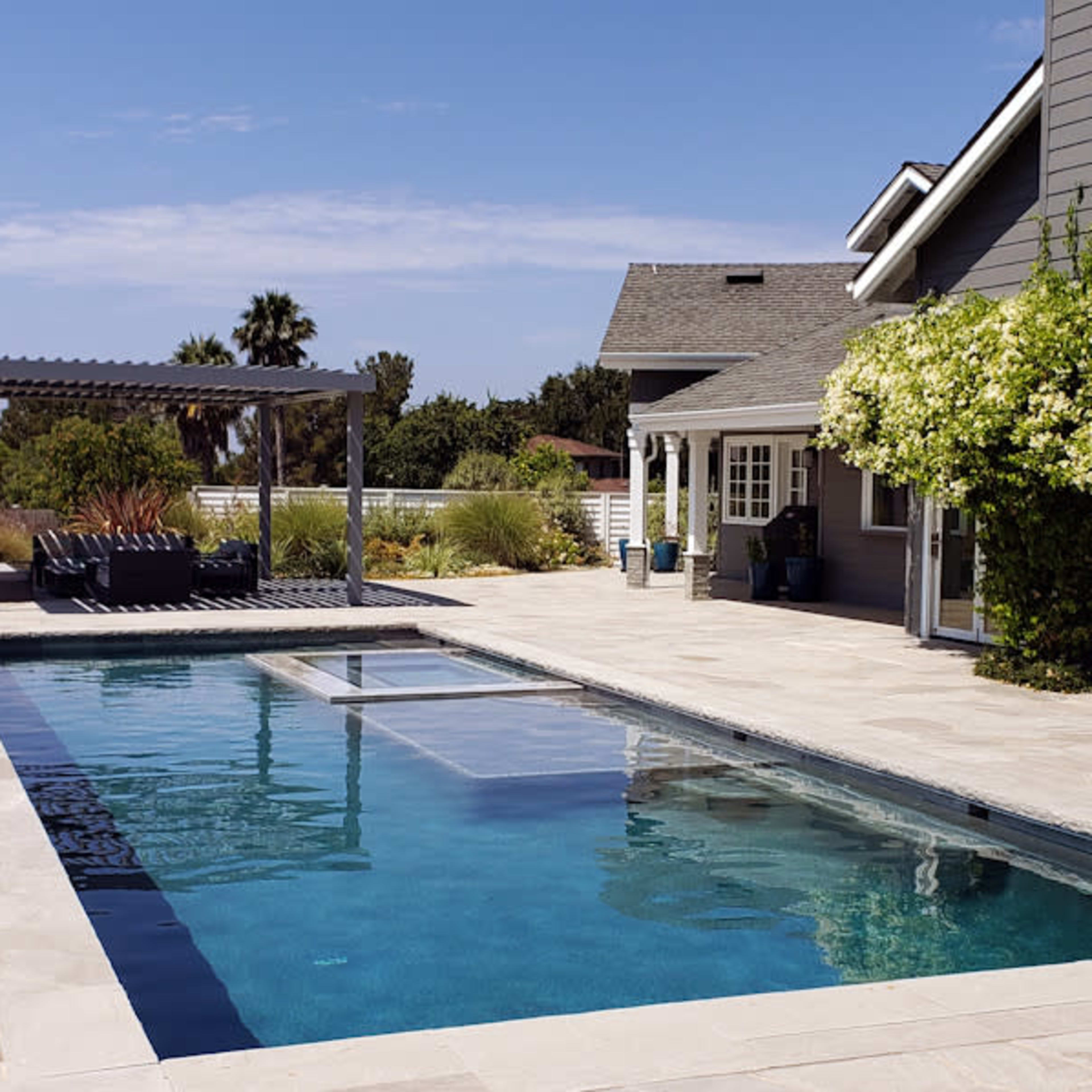 A rectangular swimming pool surrounded by a stone patio and a house with a grey exterior and flowering plants.