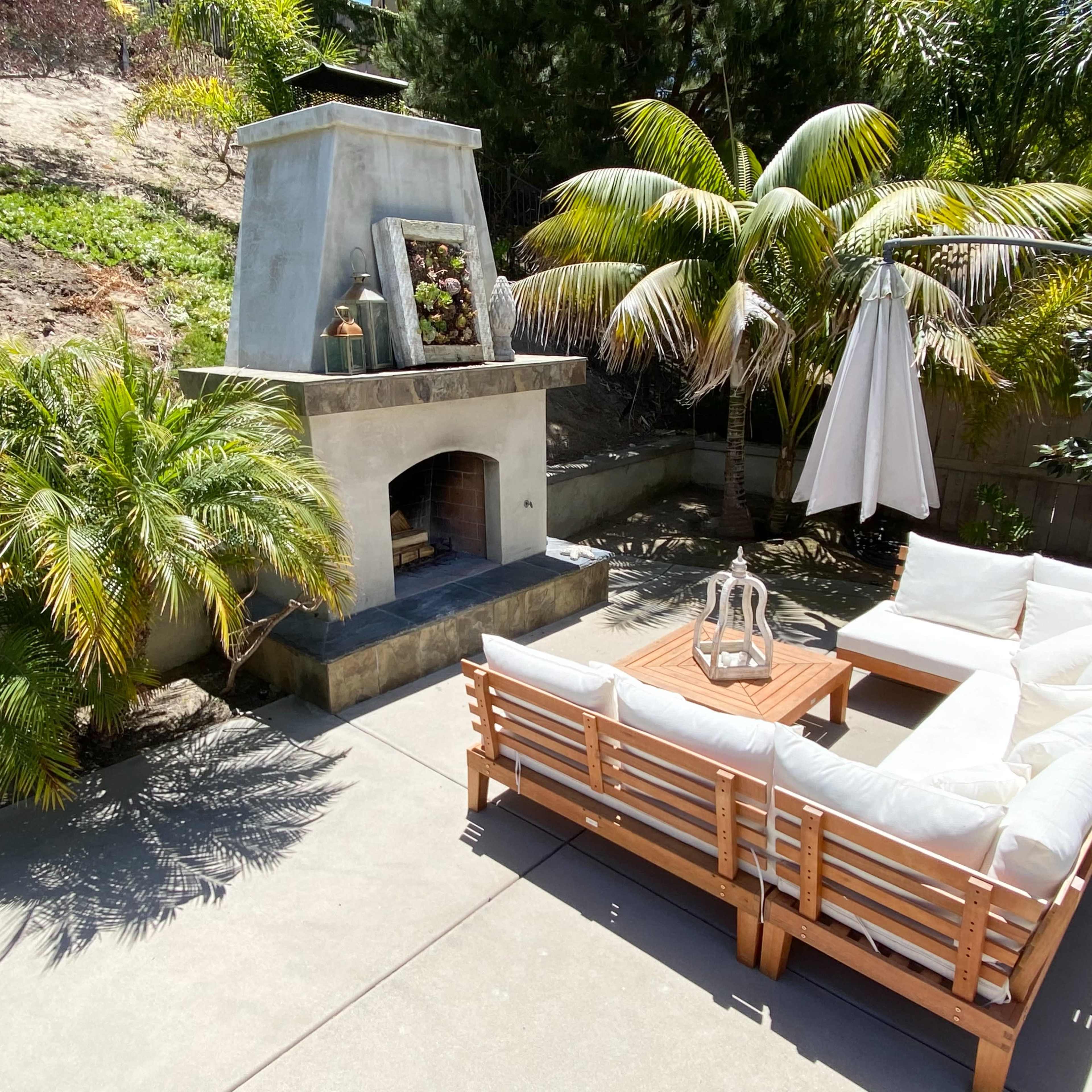 A patio area with a sectional sofa, a coffee table, an outdoor fireplace, and a large umbrella next to tropical plants and trees.