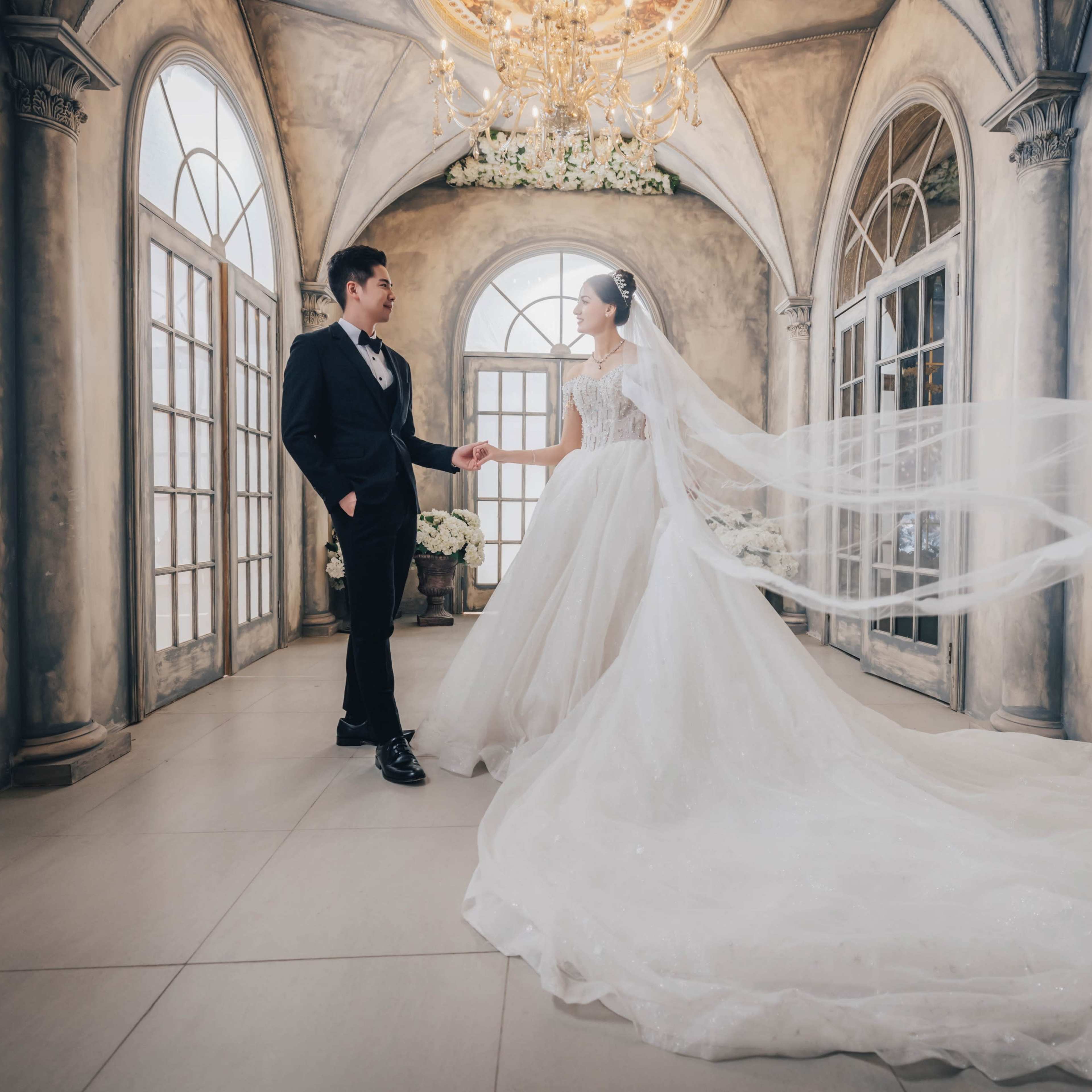 A bride in a flowing gown and a groom in formal attire stand together in an elegantly decorated indoor space with large windows and a chandelier.