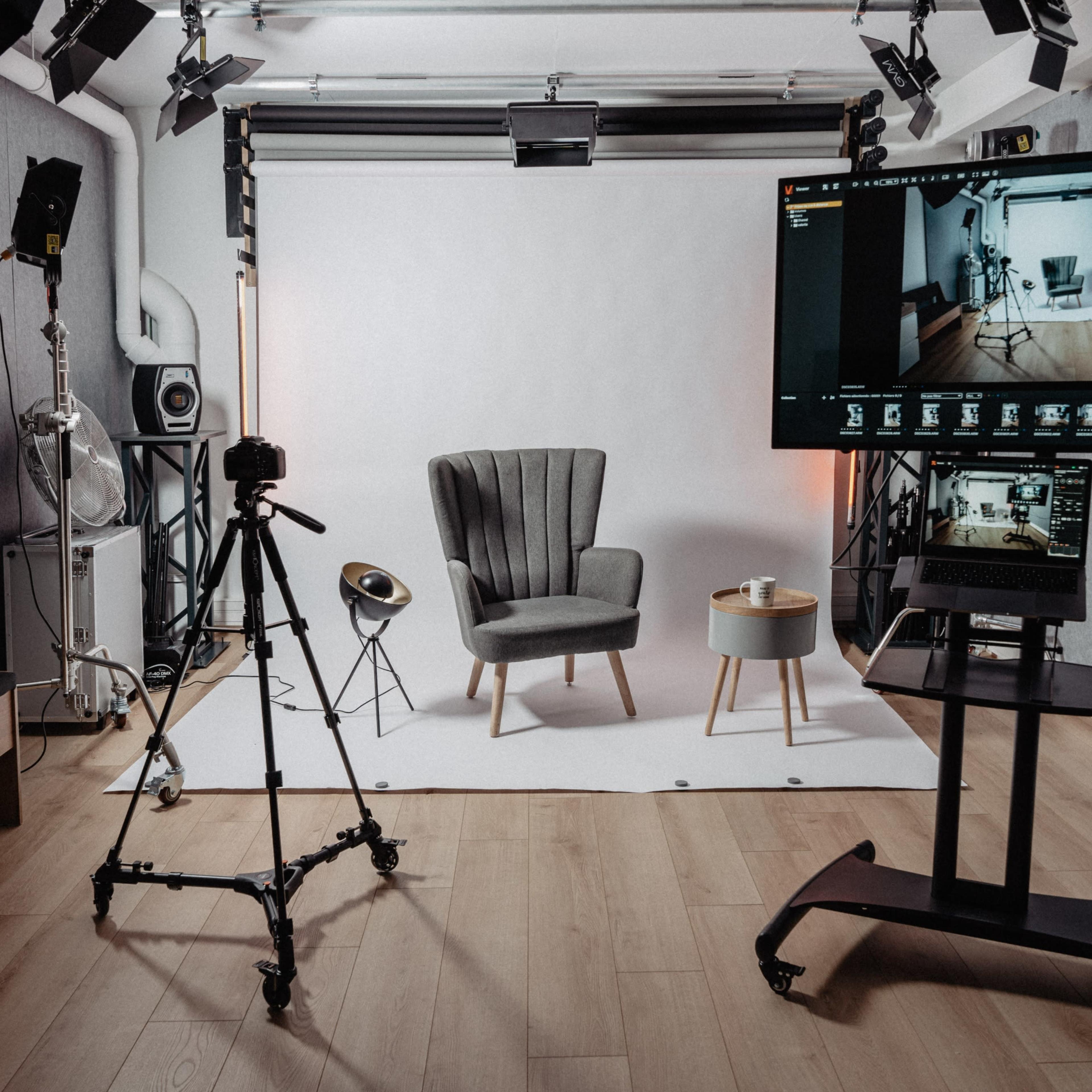 A photography studio equipped with a tripod, lighting setup, a gray armchair, a small table, and two screens displaying camera settings.