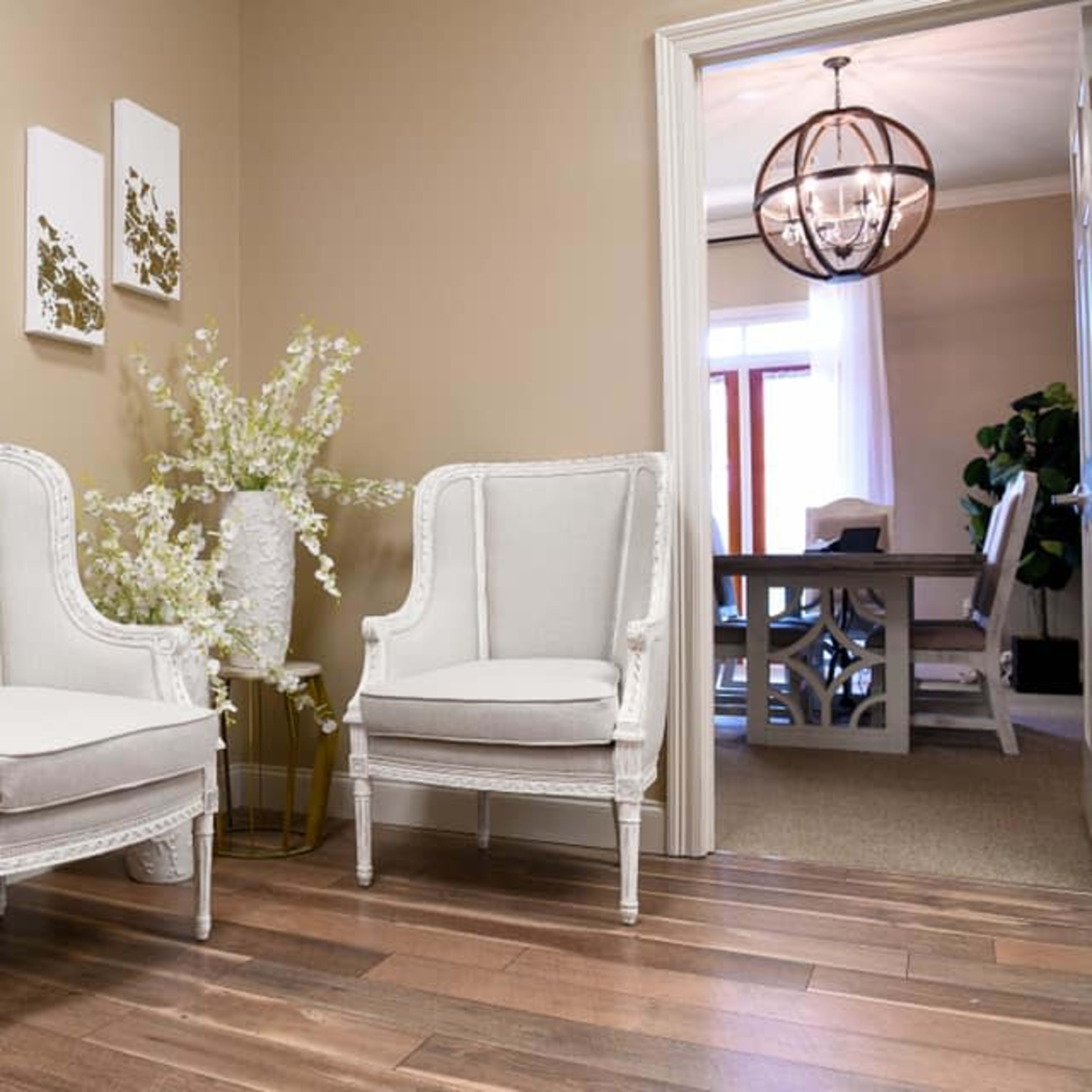 A small entryway with two white upholstered chairs flanking a tall vase filled with white flowers, and a doorway leading into a dining area.