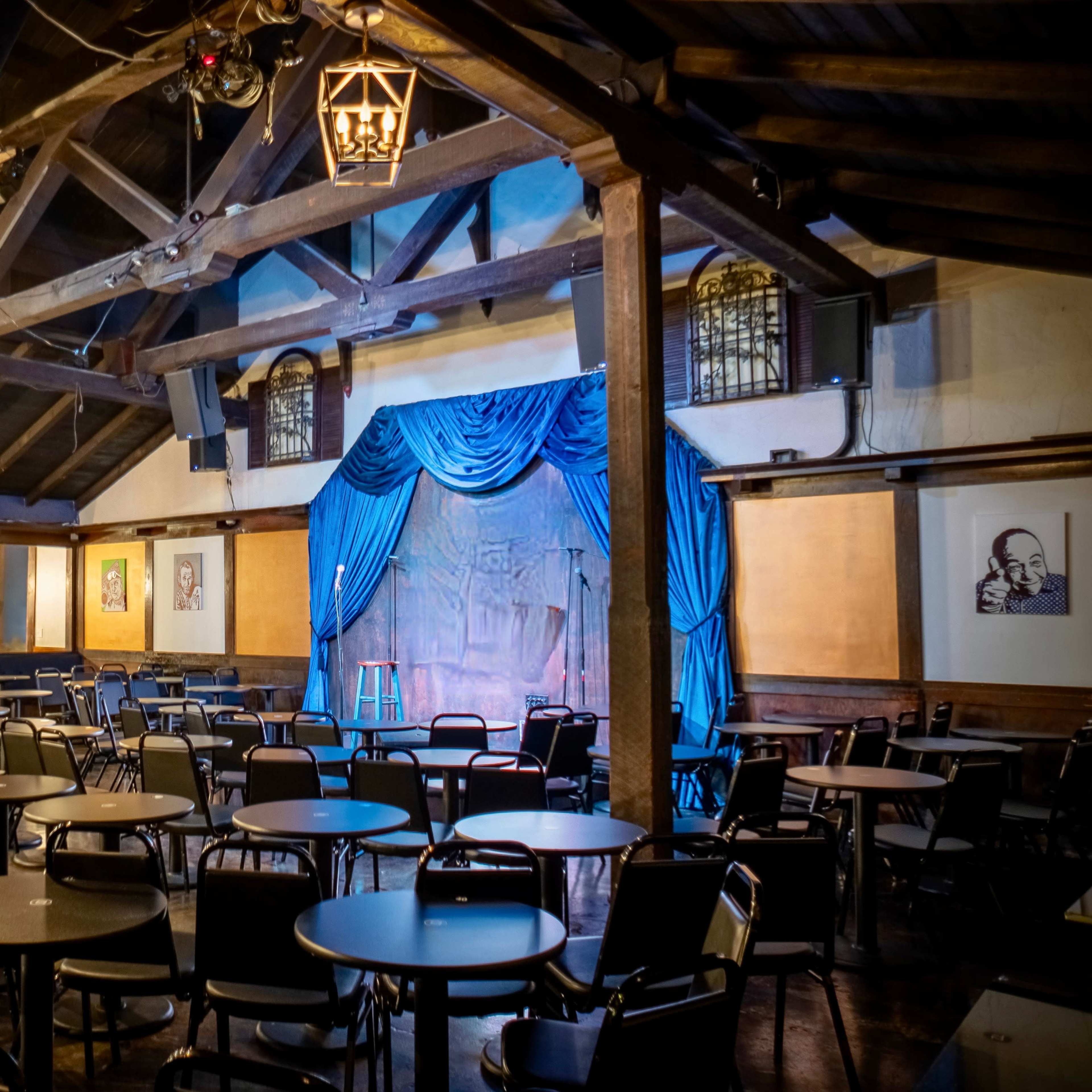 A dimly lit comedy club features a stage with blue drapery and empty tables arranged in rows around the room.