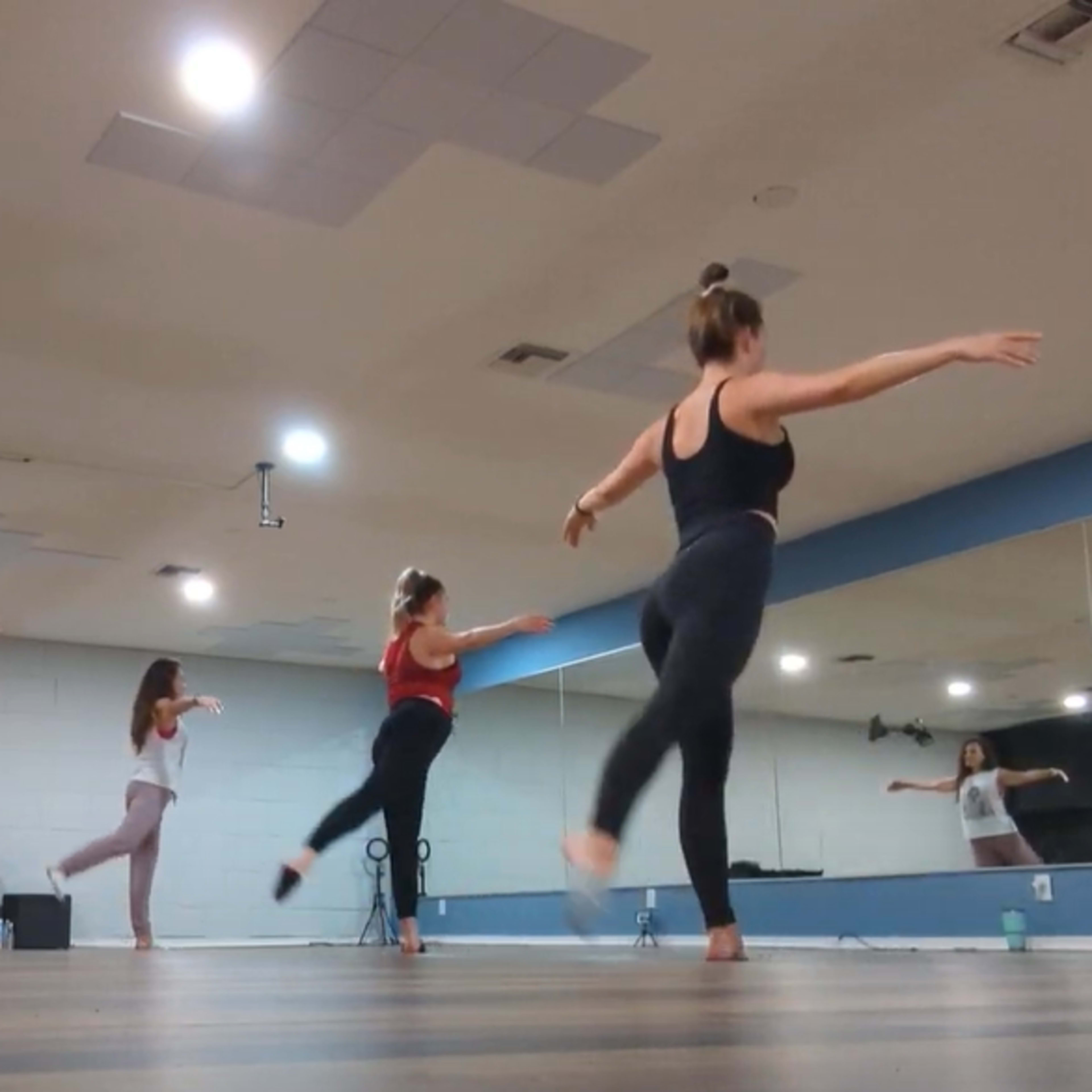 A group of dancers practices in a brightly lit studio with mirrored walls and wooden flooring.