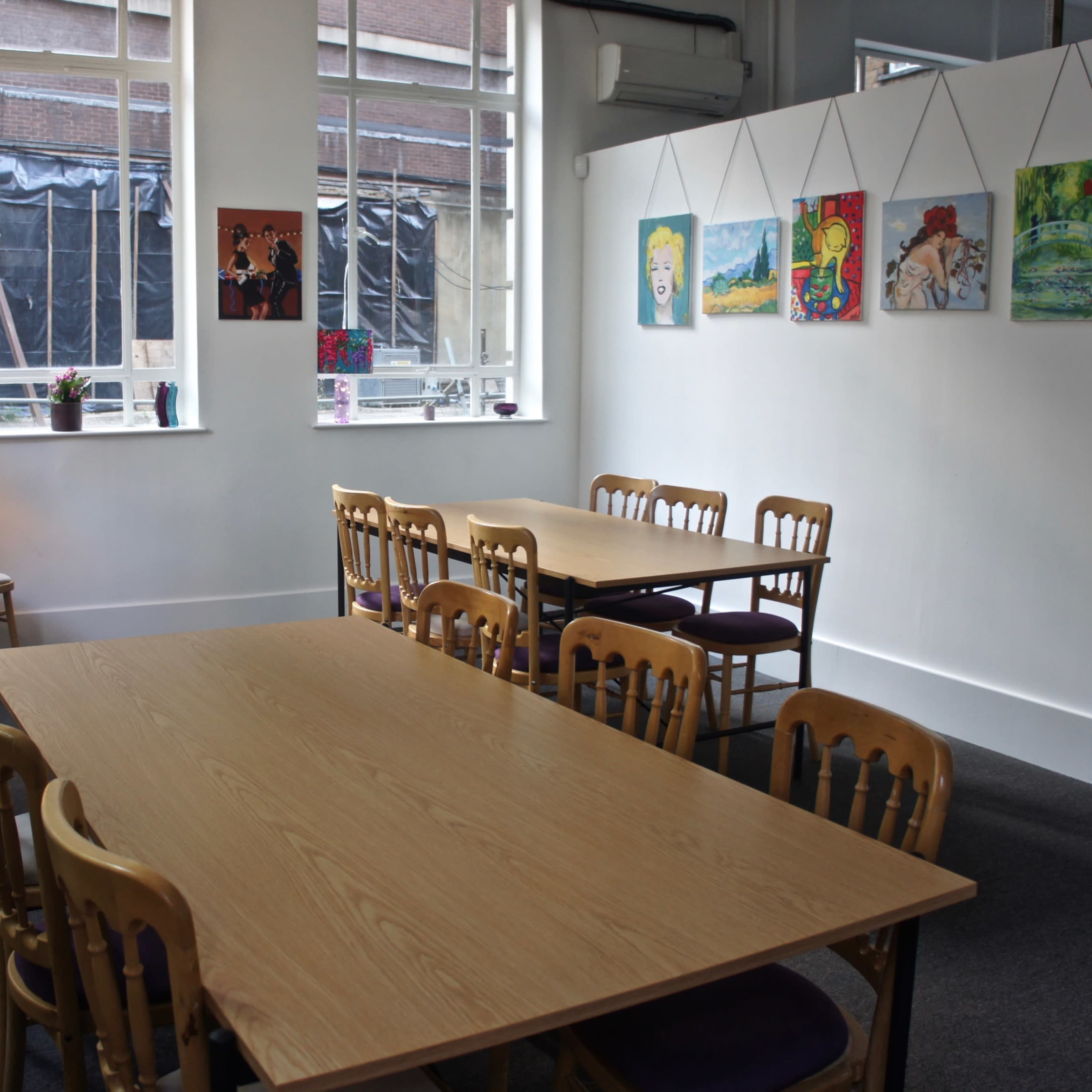 A bright, spacious room with two large tables surrounded by wooden chairs and framed artwork on the walls.
