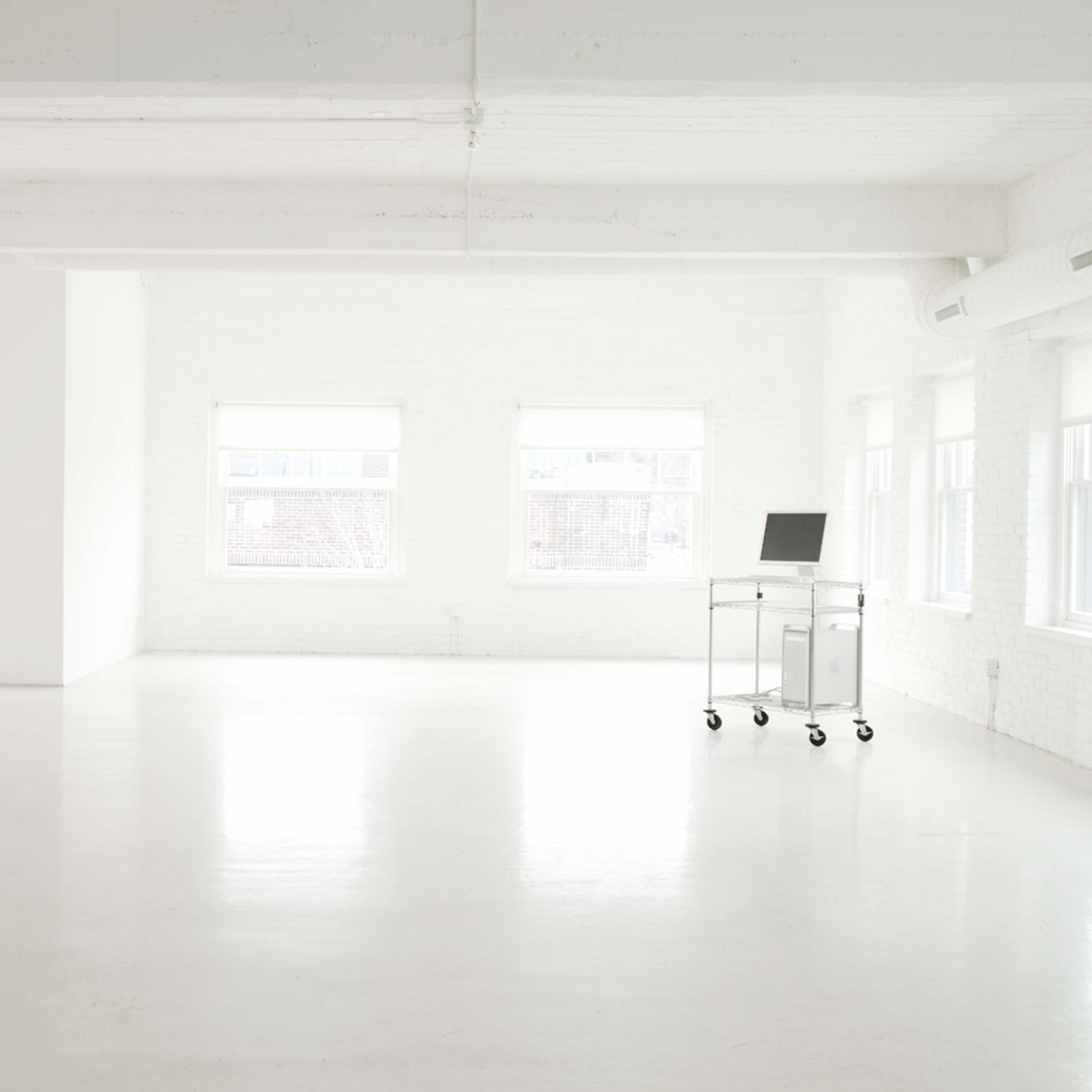 A spacious, minimalist white room with large windows, a concrete pillar, and a wheeled metal cart bearing a laptop.