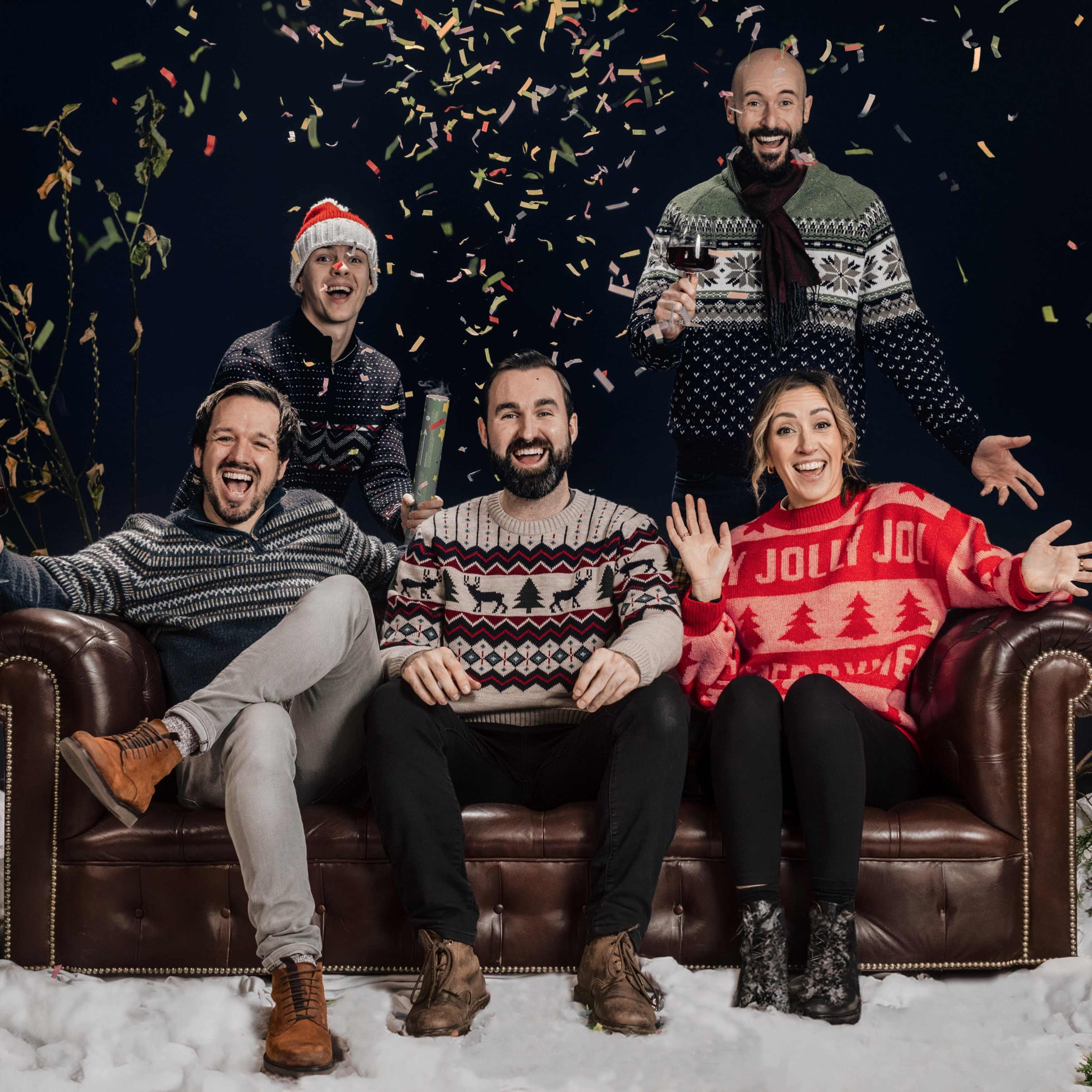 Five people wearing festive sweaters celebrate on a couch with confetti falling around them in a winter-themed setting.