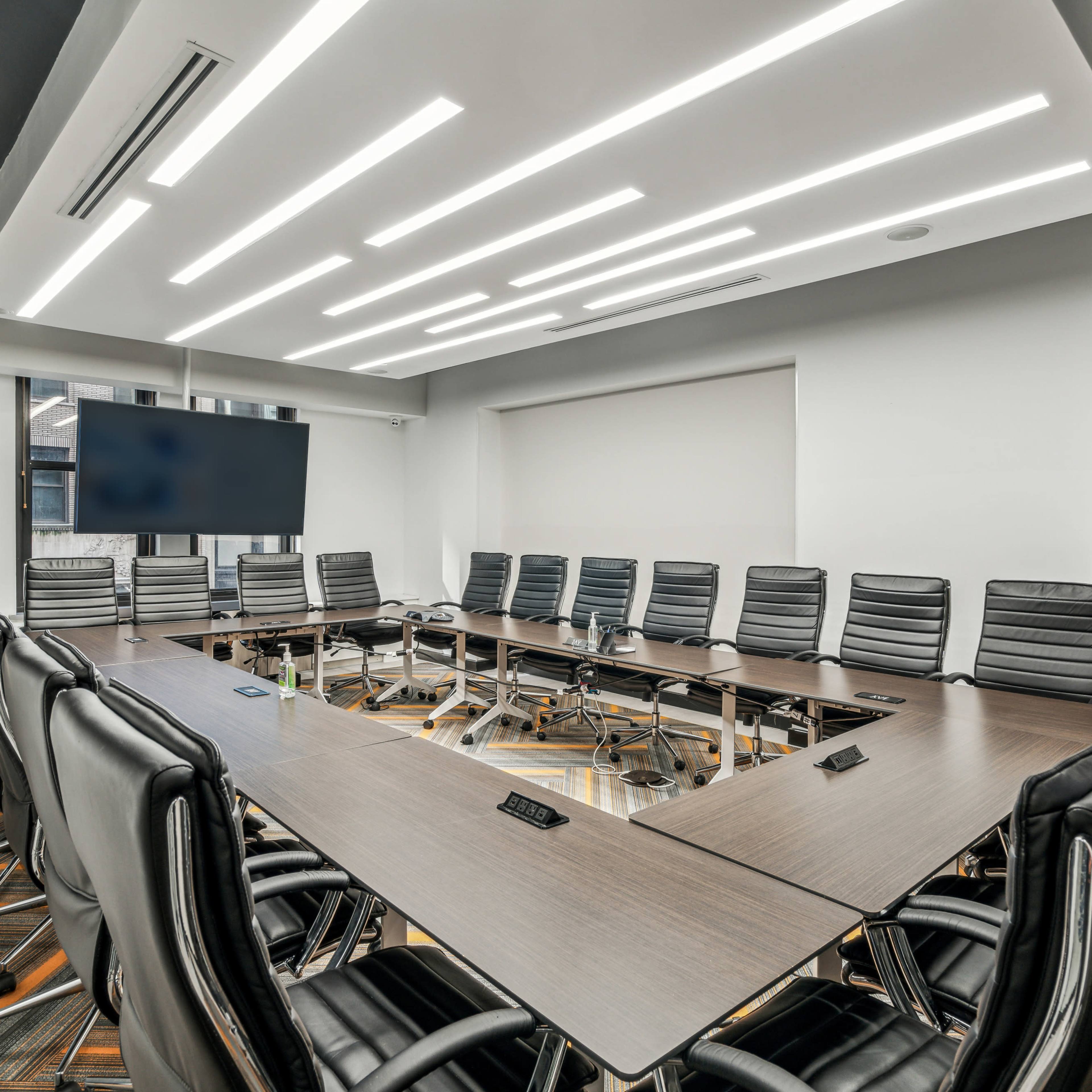 A modern conference room with a large rectangular table surrounded by black leather office chairs and a wall-mounted screen.