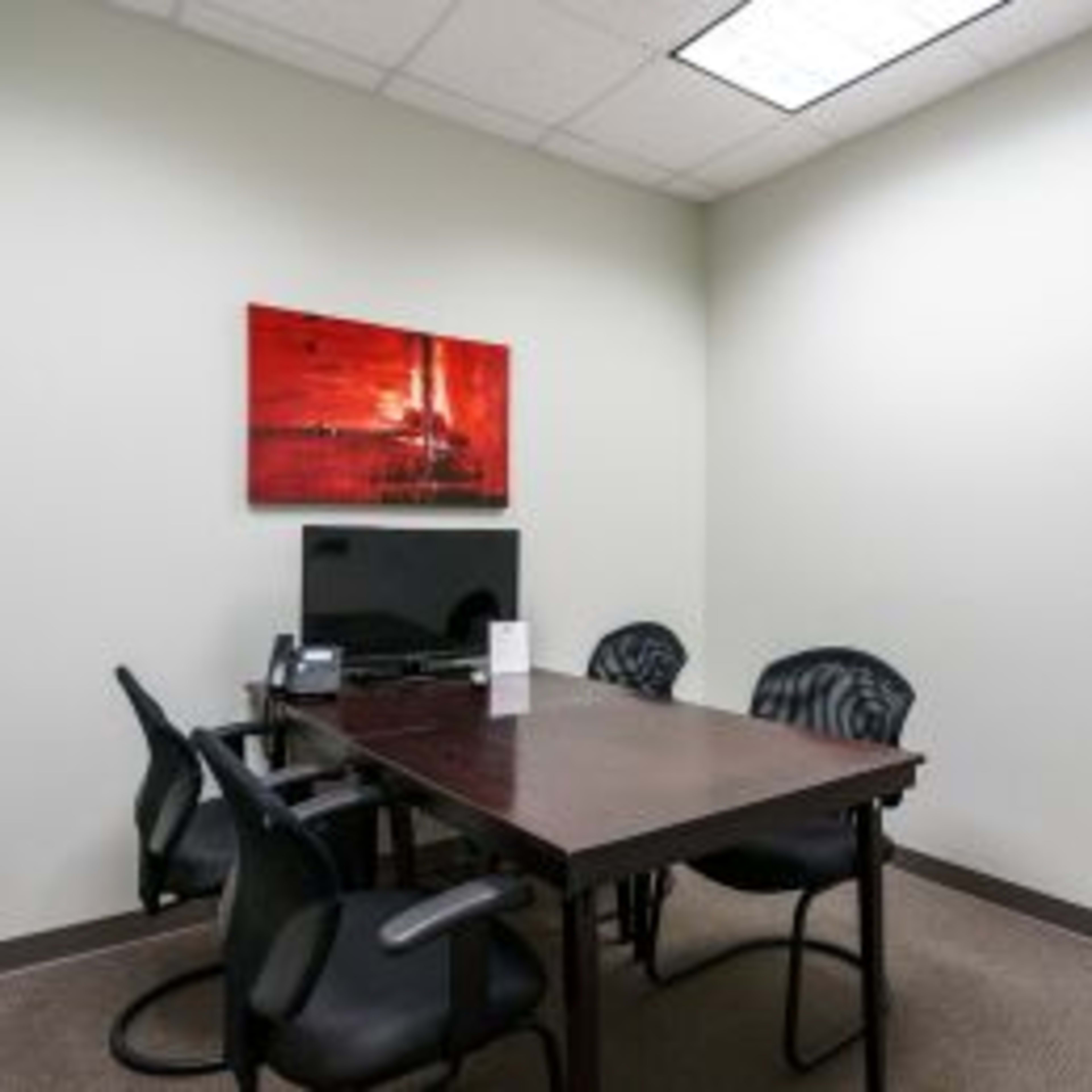 A small office room with a rectangular table, four black chairs, a television, and a piece of artwork on the wall.
