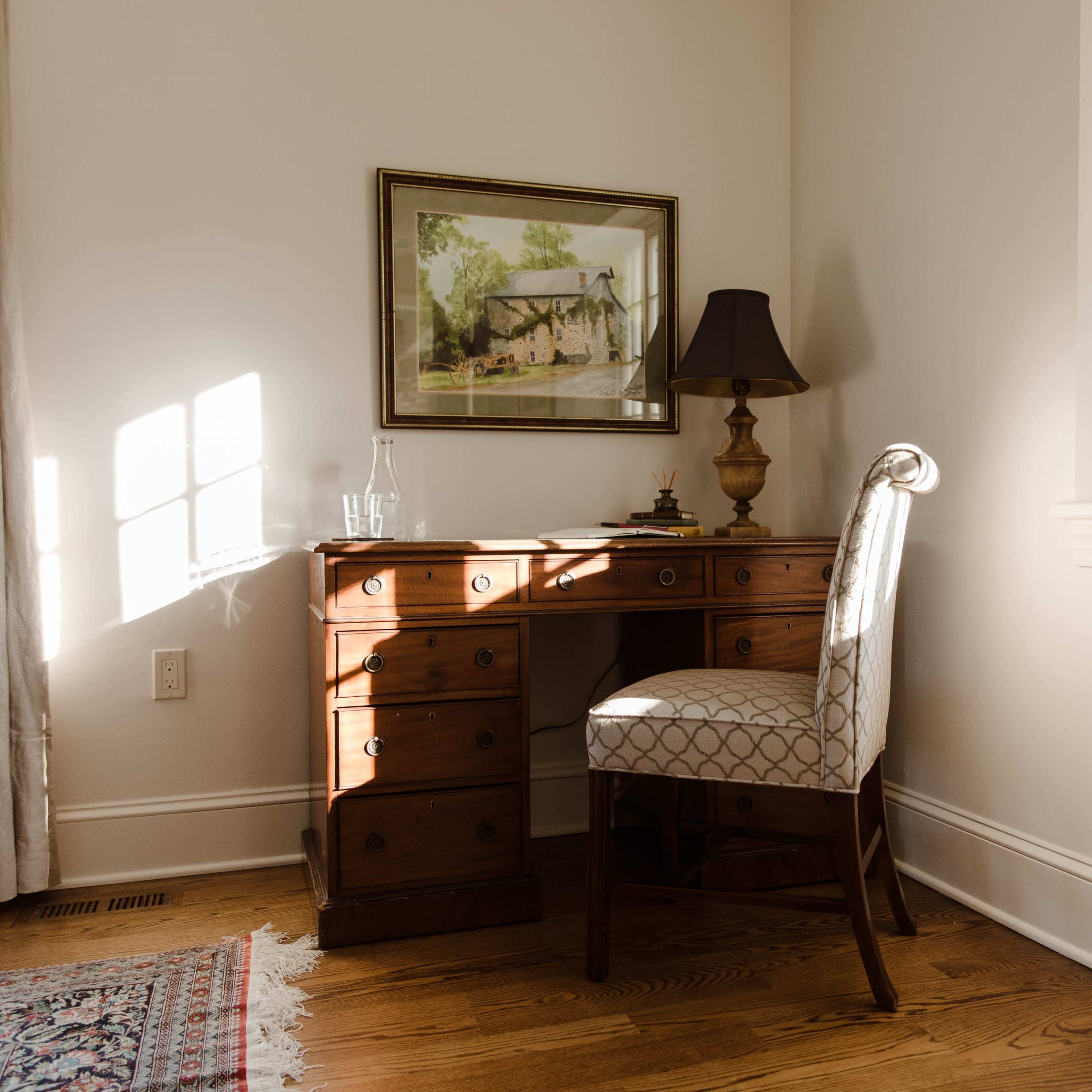 A wooden desk with a matching chair and a lamp is positioned beside a window in a well-lit room.