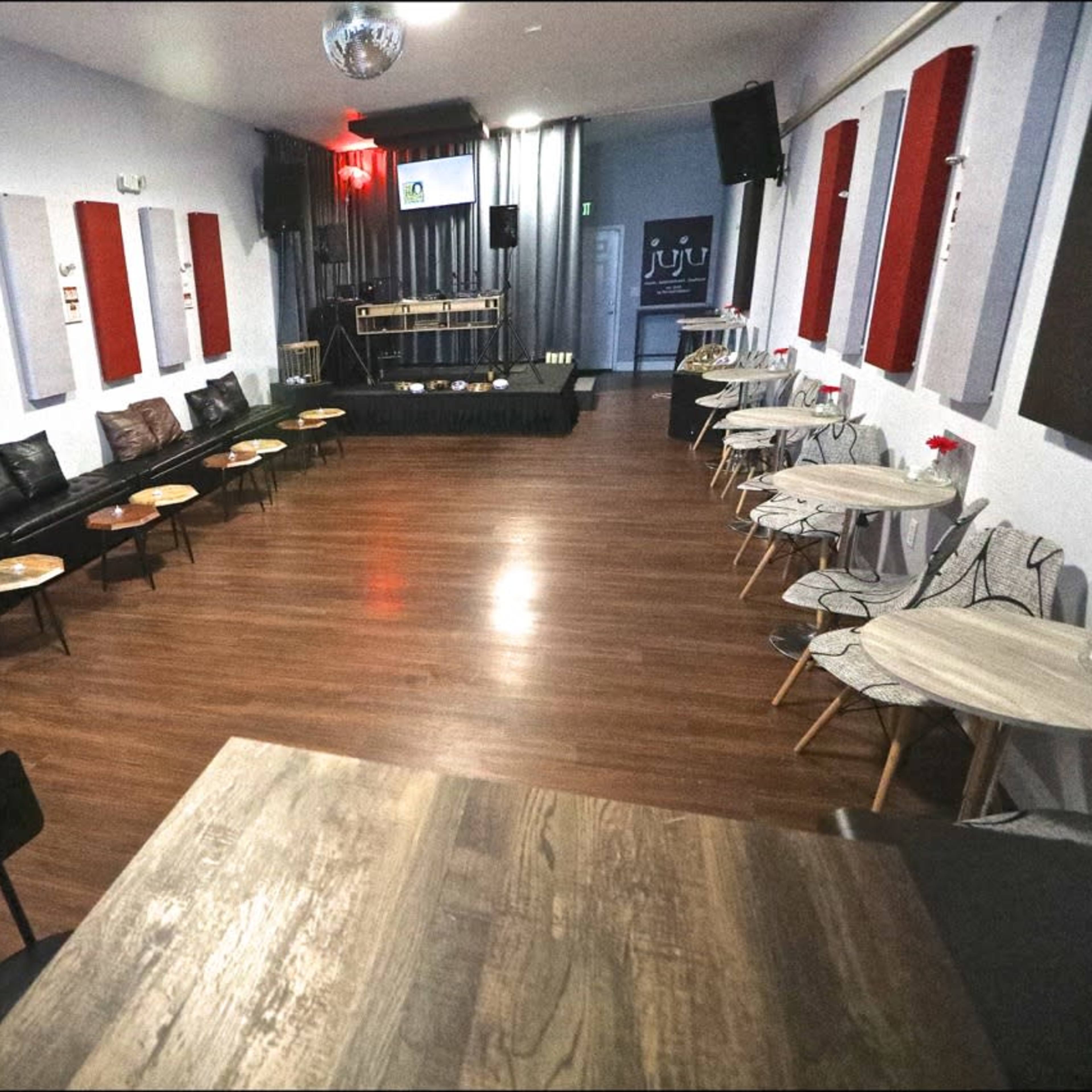 A spacious interior of a lounge or event space with black leather seating, wooden tables, and sound equipment set up at the front.