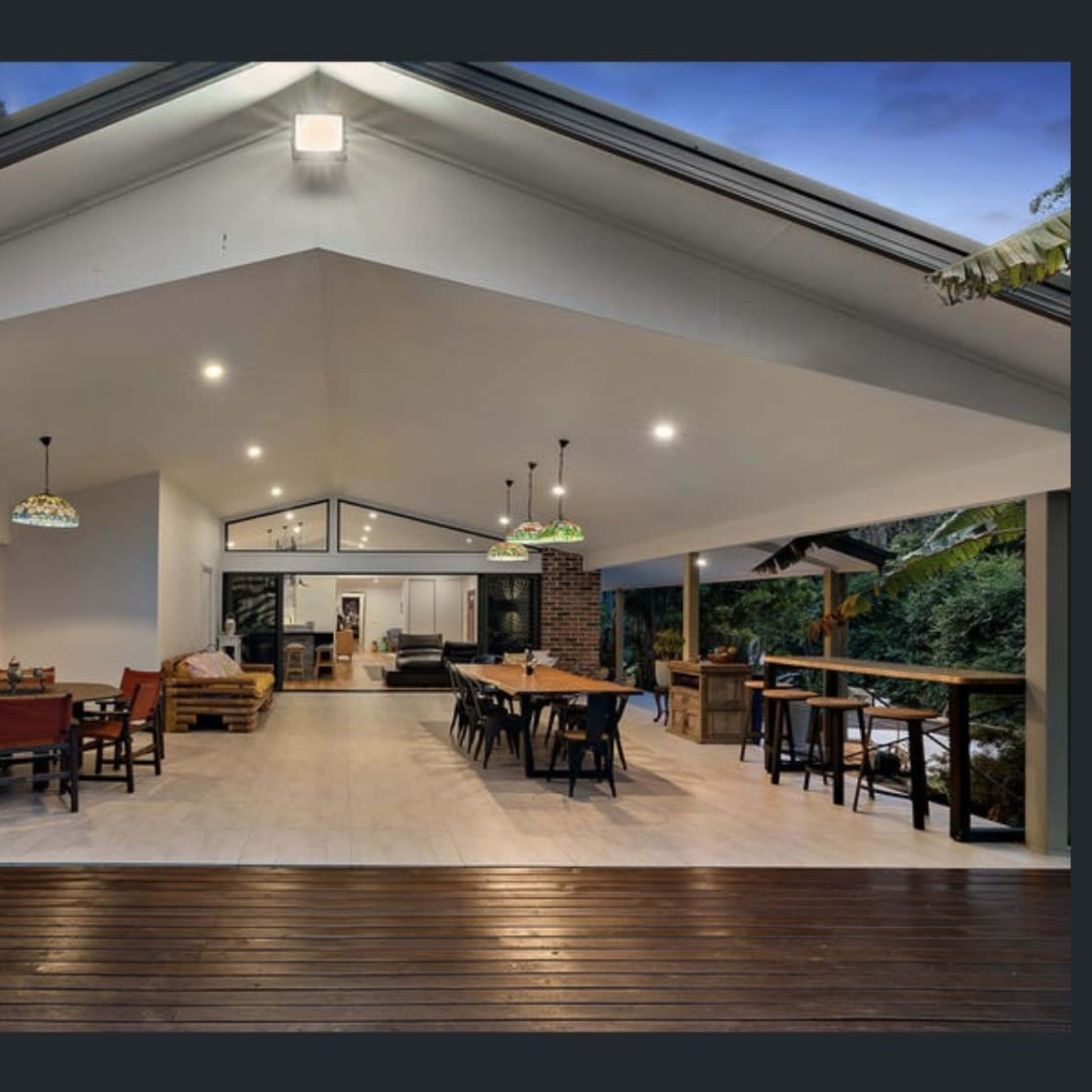 A spacious outdoor living area with wooden flooring, featuring dining tables and chairs under pendant lights, connected to a modern interior space.