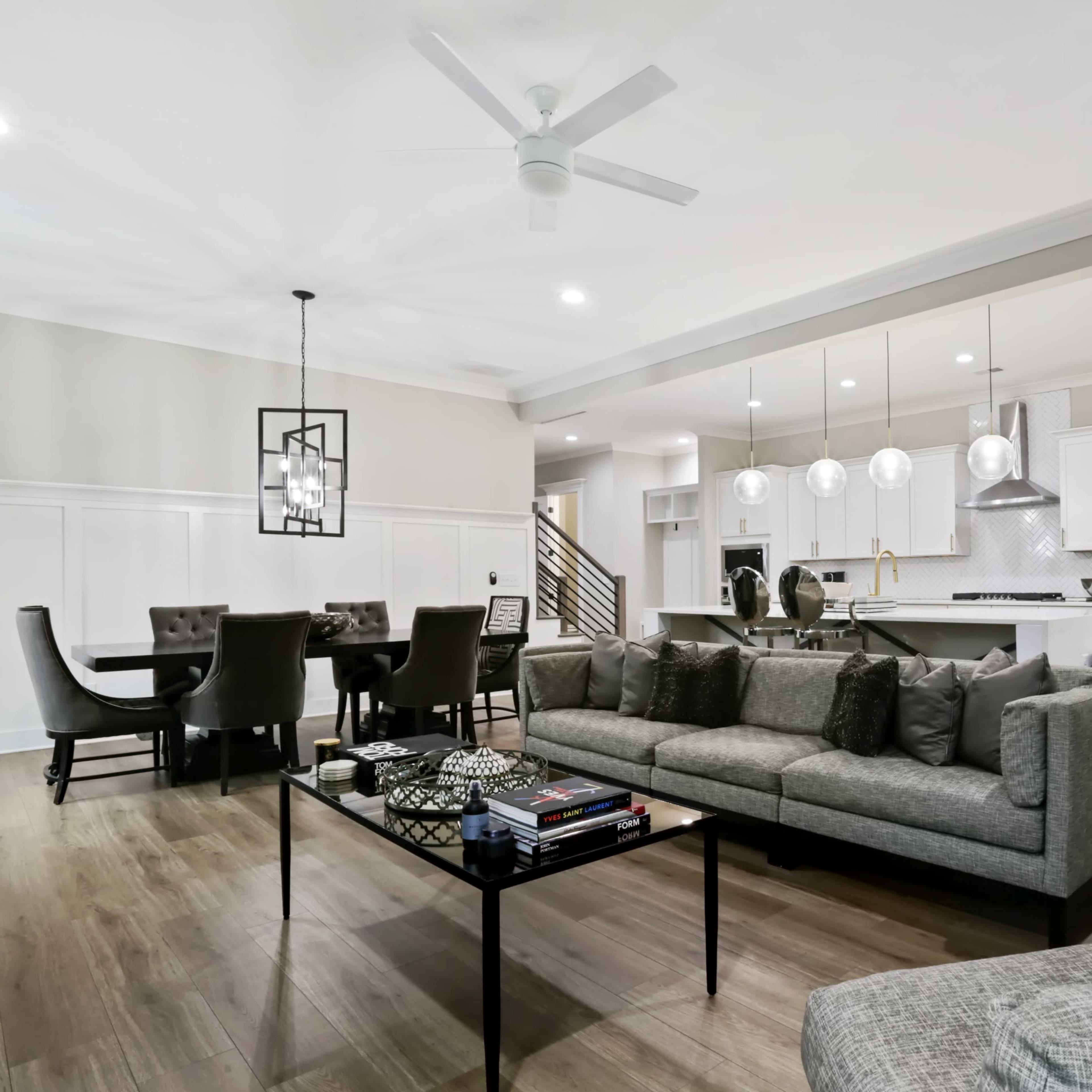 A modern open-concept living space featuring a living area with a gray sectional sofa, a dining table with black chairs, and a kitchen with stainless steel appliances and pendant lighting.