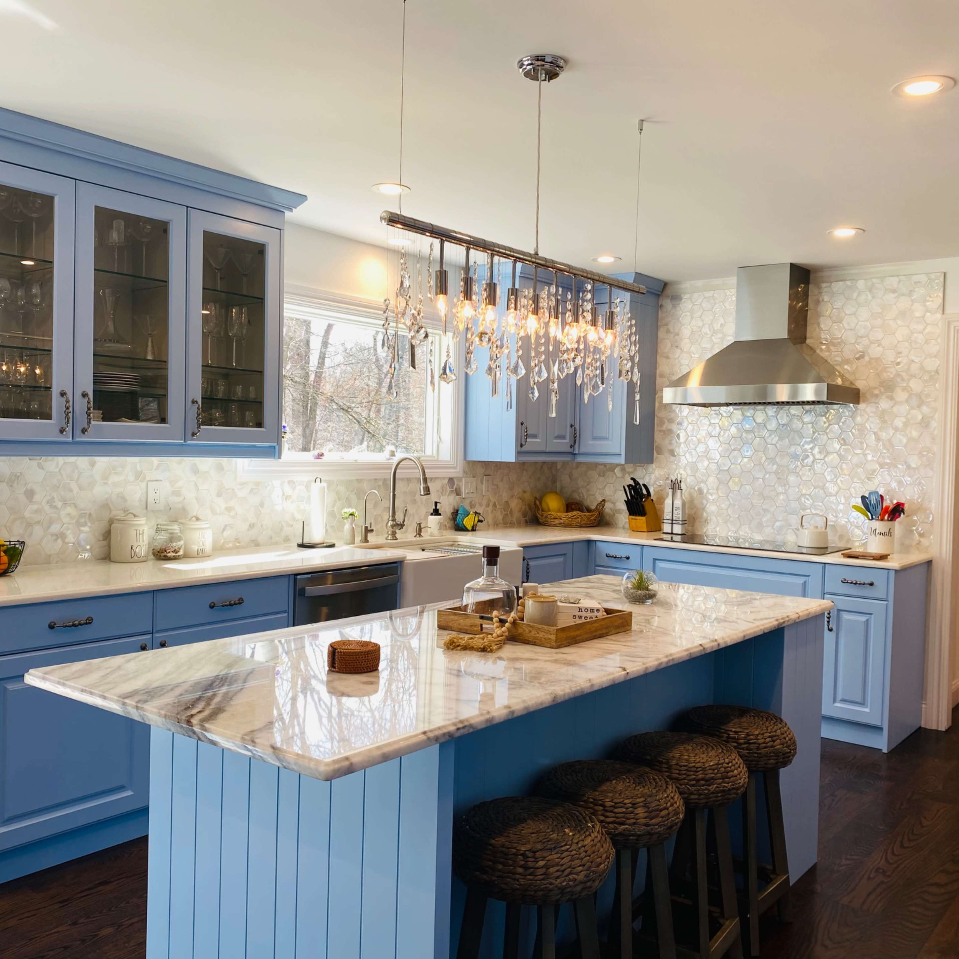The kitchen features blue cabinetry, a marble island with bar stools, and a chandelier above, with a modern stainless steel range and backsplash.