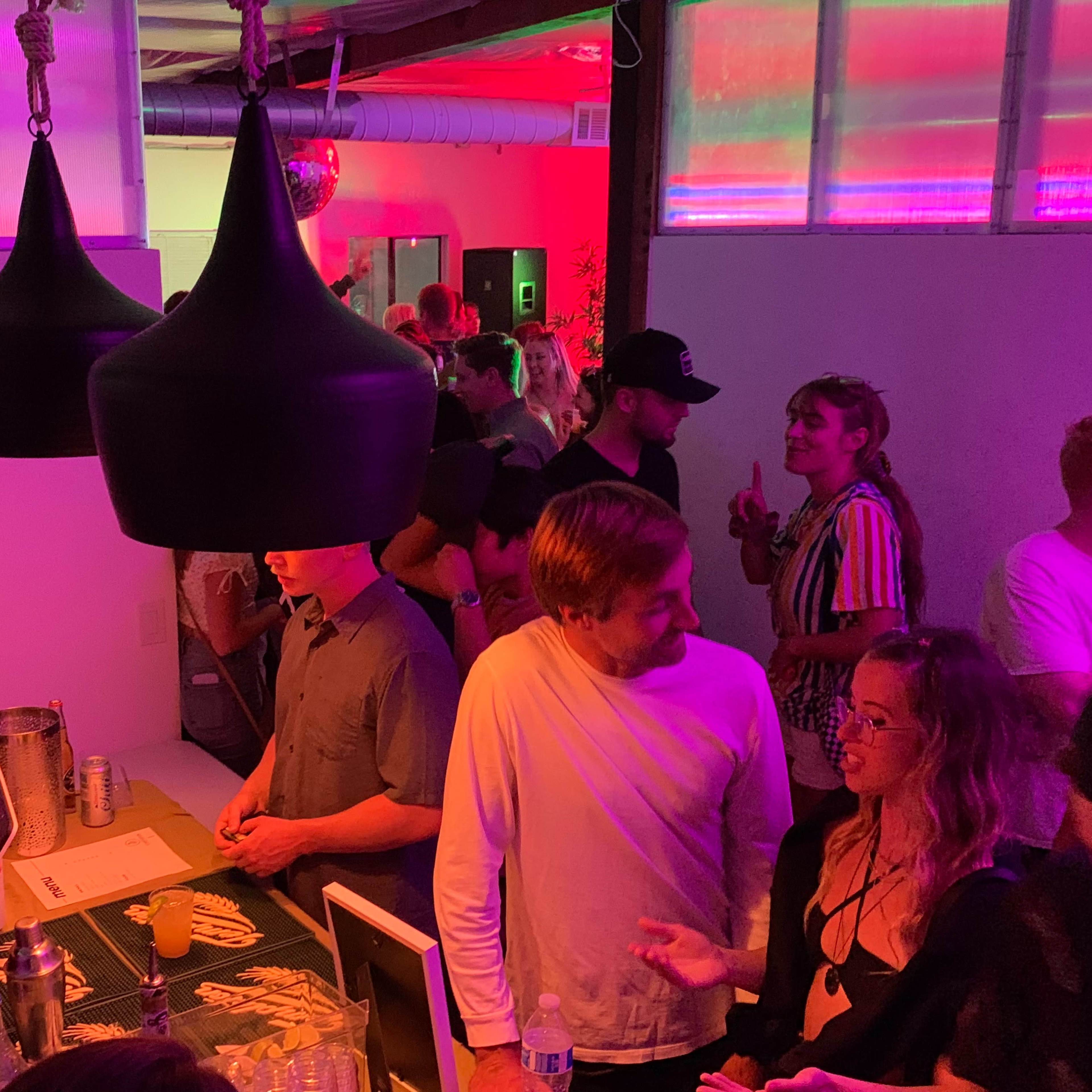 A crowd socializes in a dimly lit room with colorful lighting, while two individuals chat near a bar area.