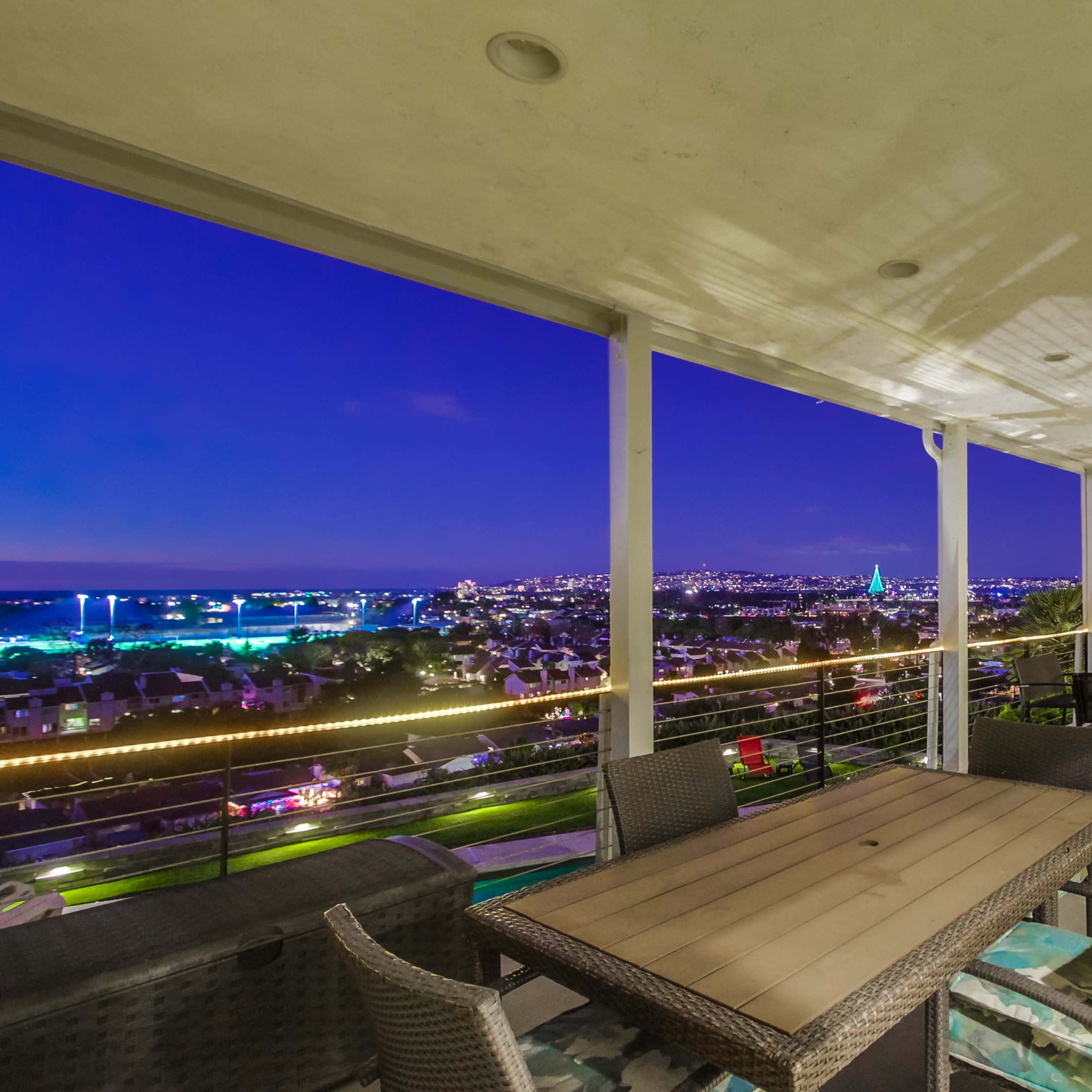 A spacious balcony with a wooden dining table and chairs, overlooking a city skyline illuminated at night.