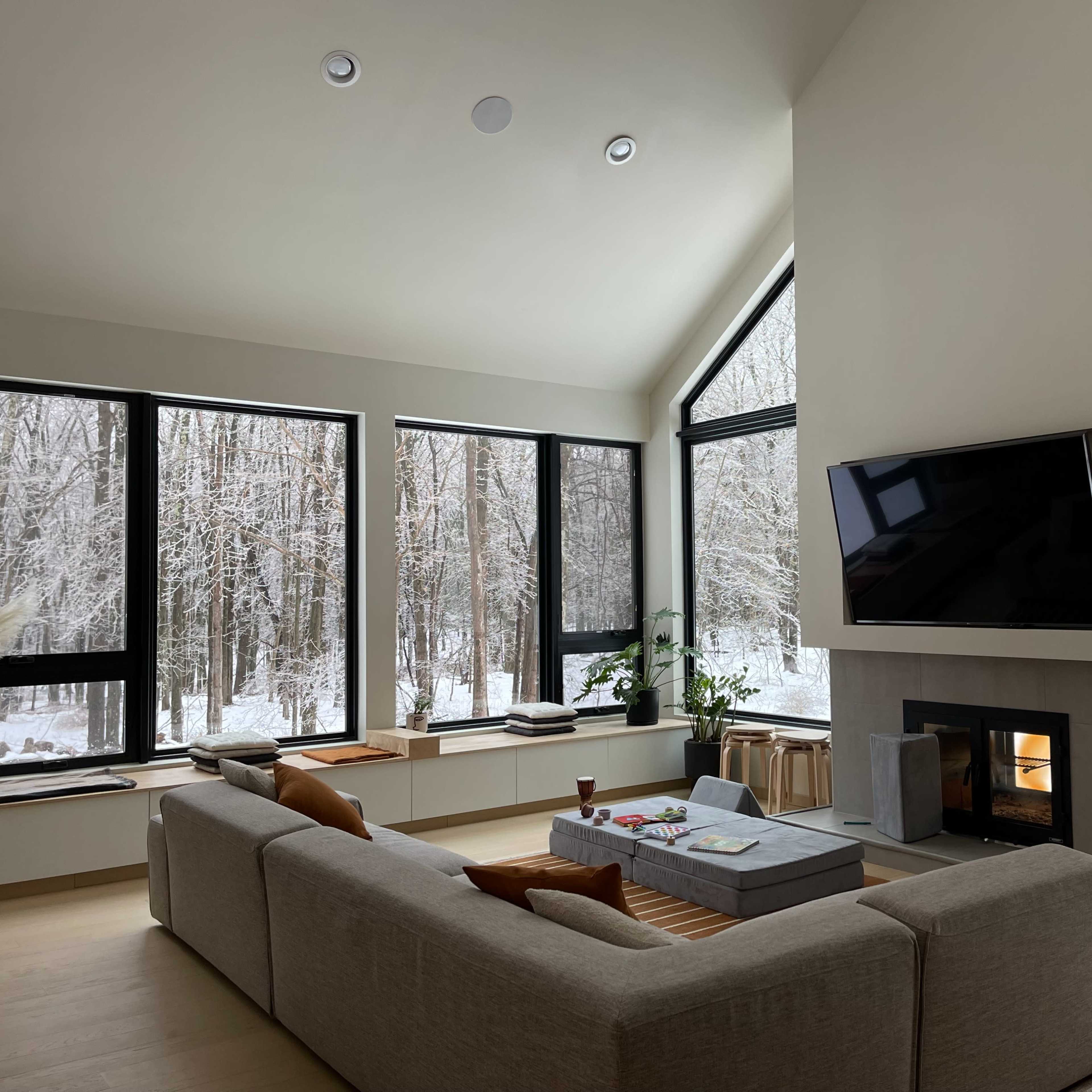 A modern living room features large windows overlooking a snowy forest, with a sectional sofa, a coffee table, and a wall-mounted television above a fireplace.