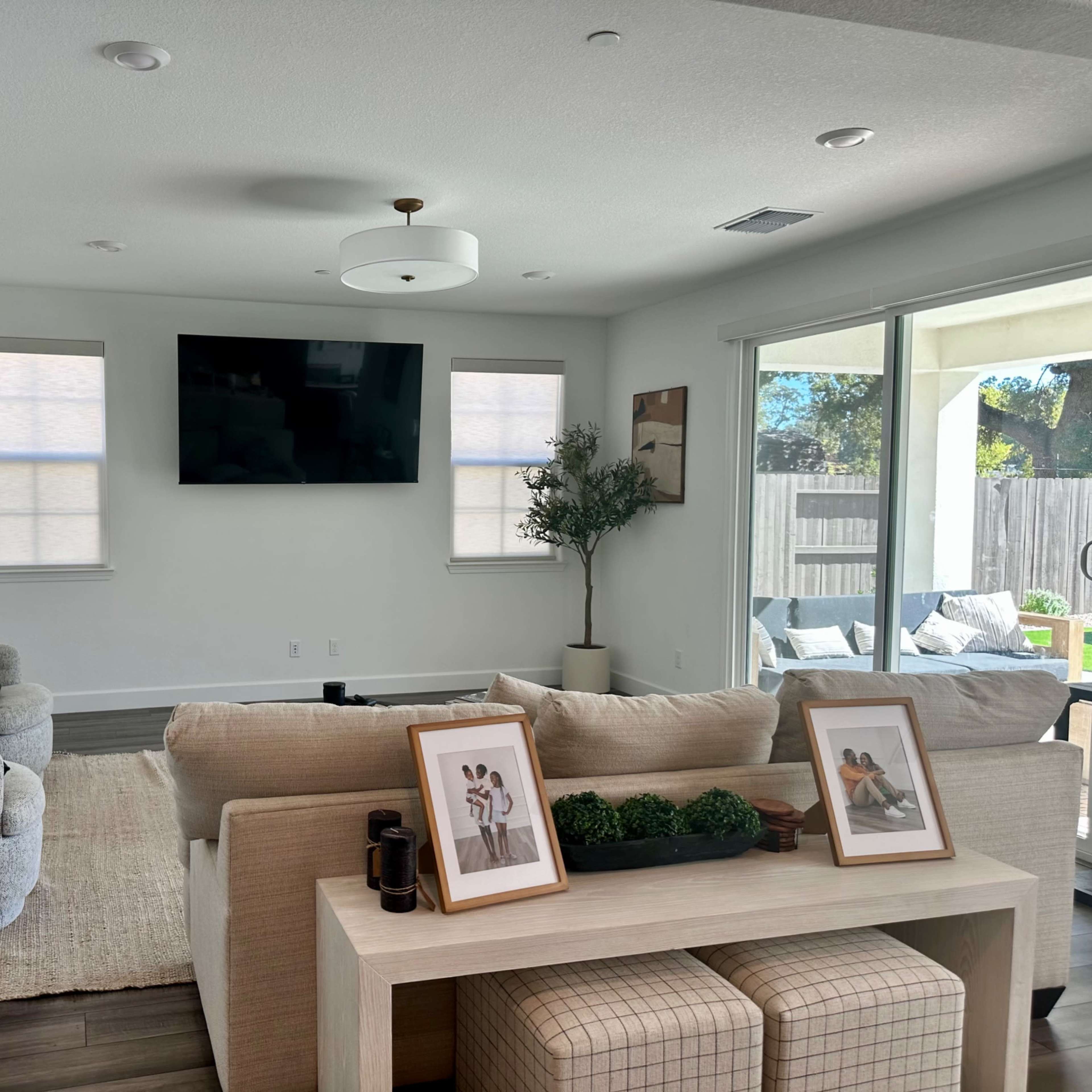 A modern living room features a neutral color palette, a large wall-mounted television, comfortable seating, and a sliding glass door leading to an outdoor area.