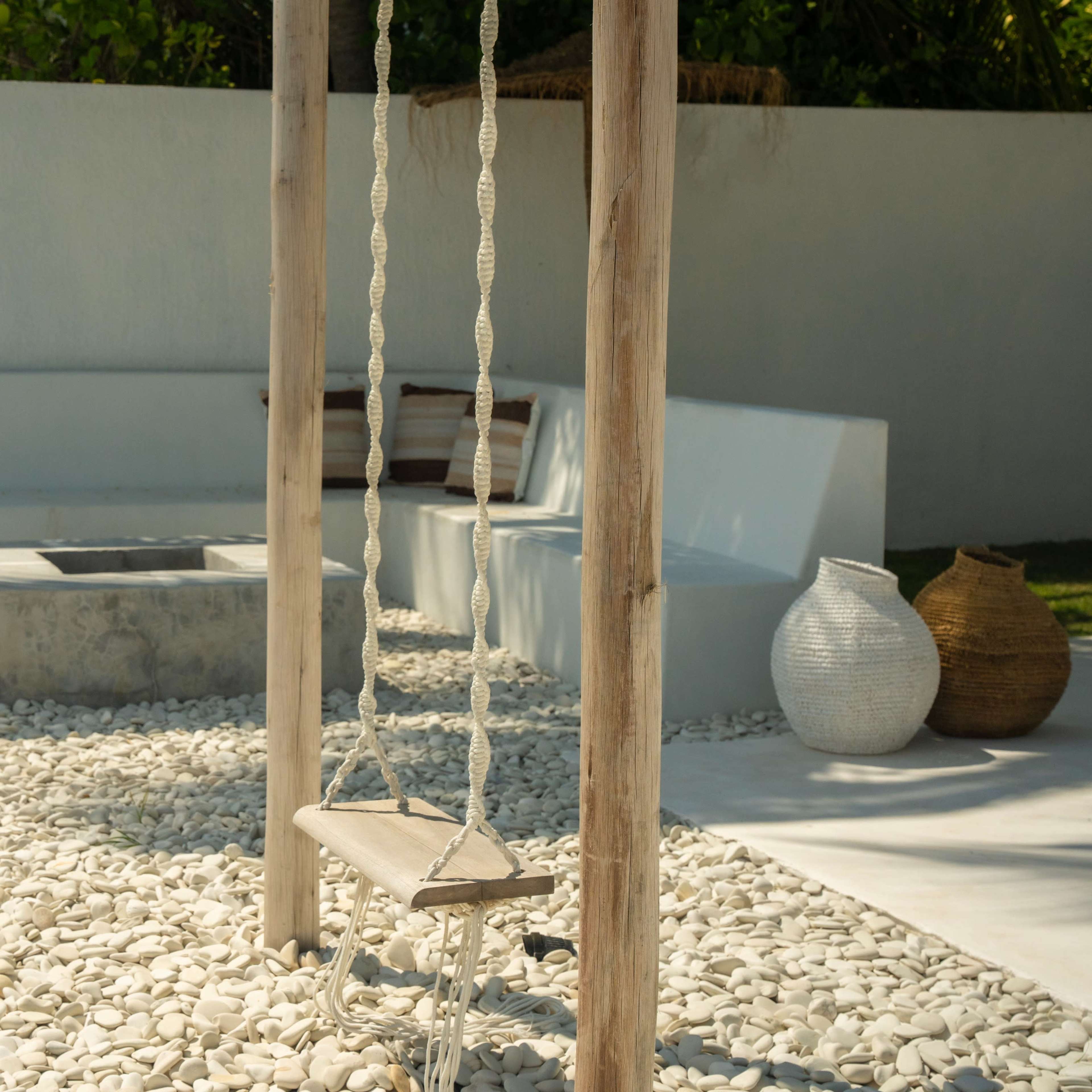 A wooden swing hangs from a post in a minimalist outdoor space with smooth white stones, a concrete fire pit, and modern seating arrangements.