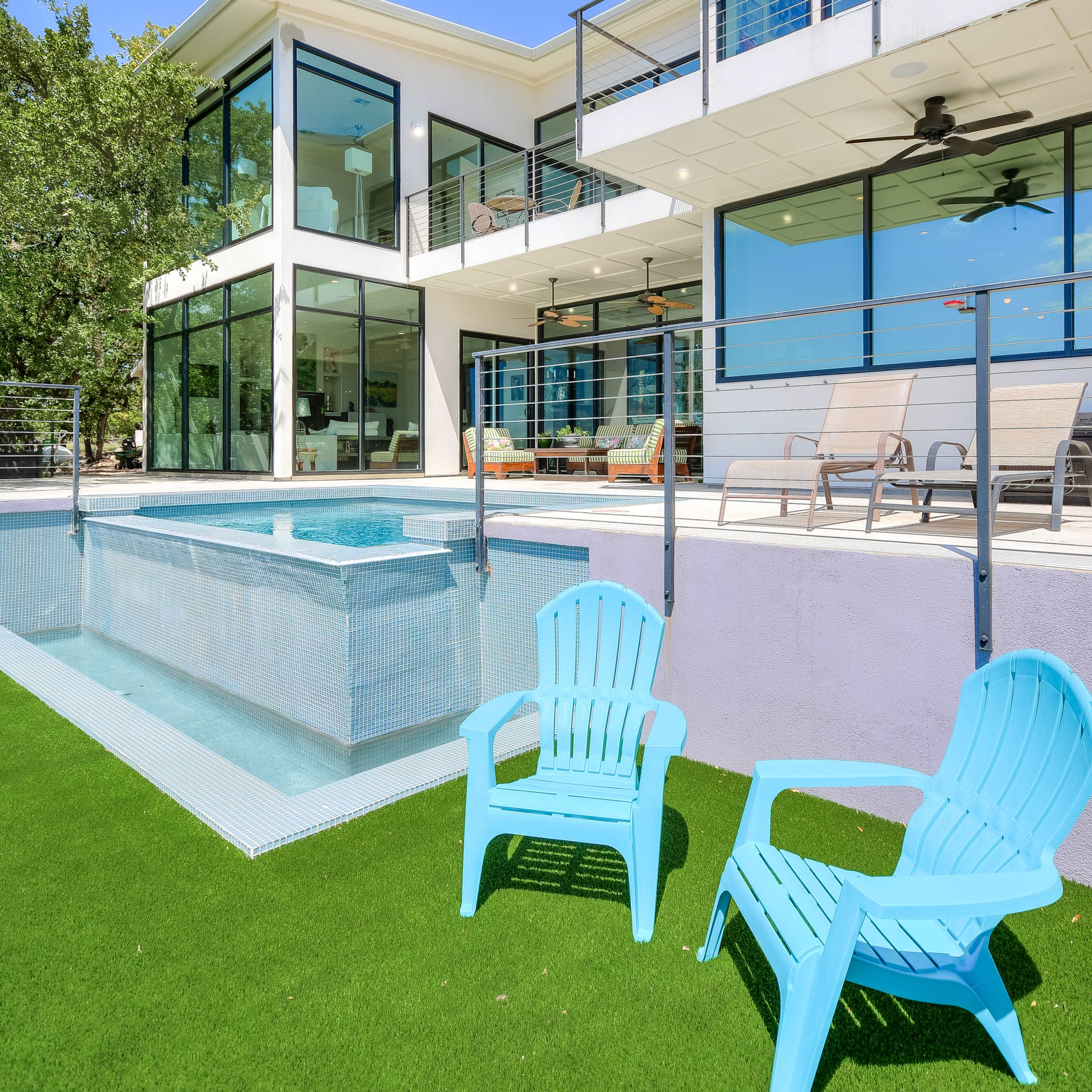 A modern house with large glass windows features a swimming pool and two blue chairs on a grassy area.