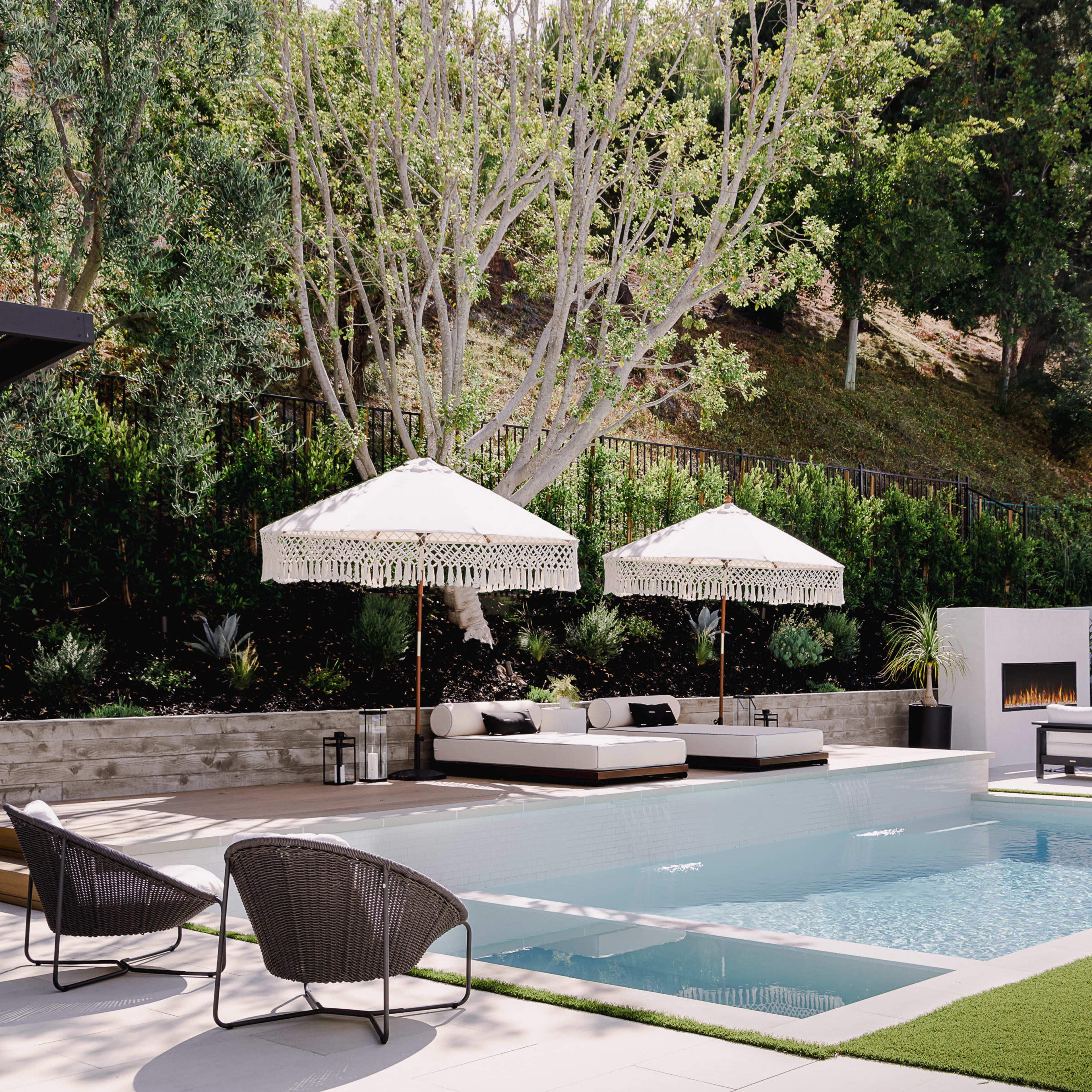 A modern pool area with two umbrellas over lounge chairs, surrounded by greenery and a fire feature on the side.