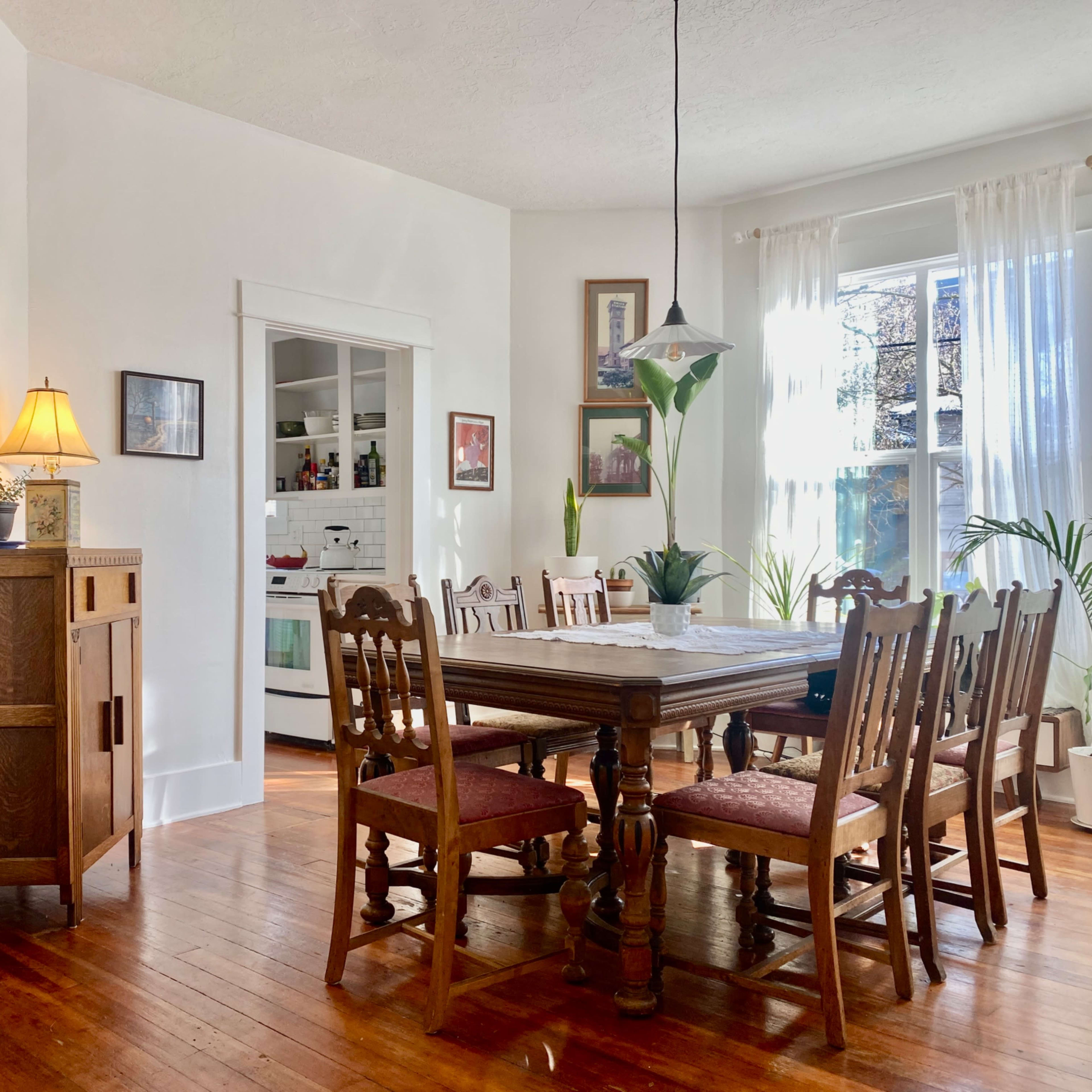 A wooden dining table with chairs is set in a bright room featuring large windows and plants, with a kitchen visible in the background.